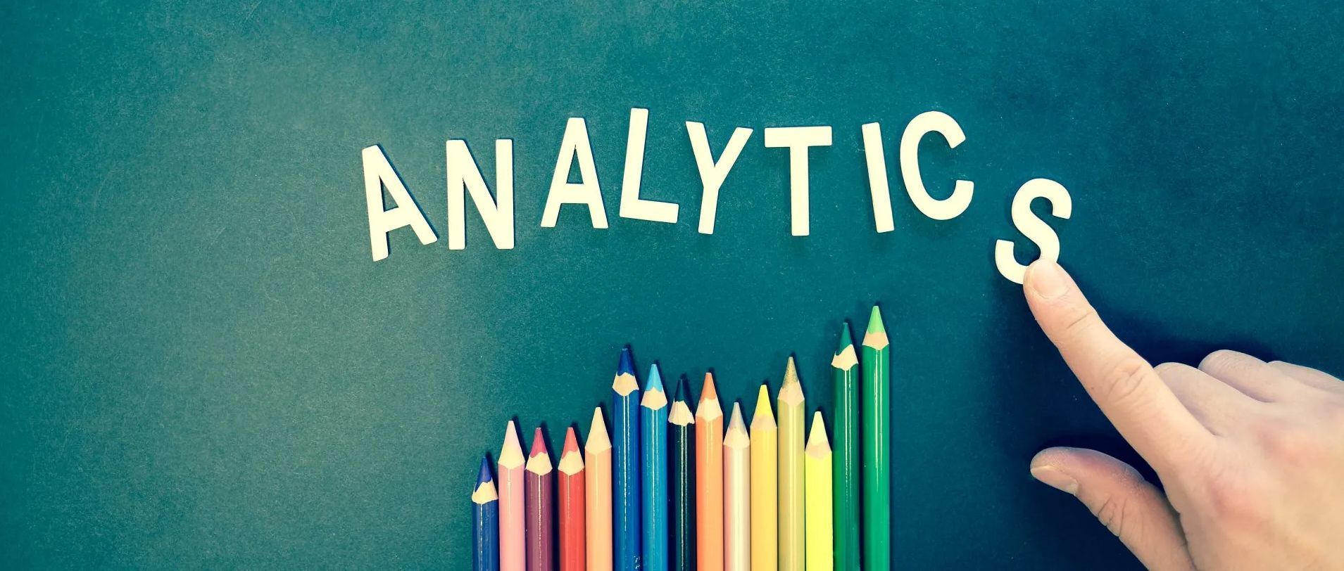 A hand places the letter S next to the word ANALYTICS spelled out in white letters above a row of colorful pencils.