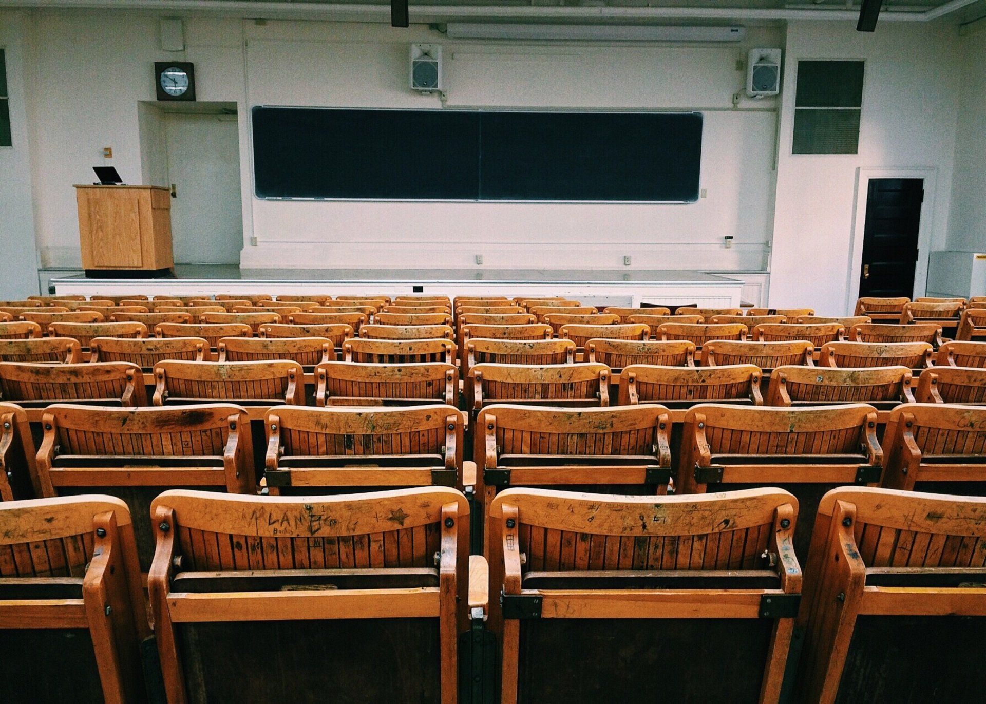 Empty lecture hall with rows of wooden chairs facing a black chalkboard and a wooden podium.