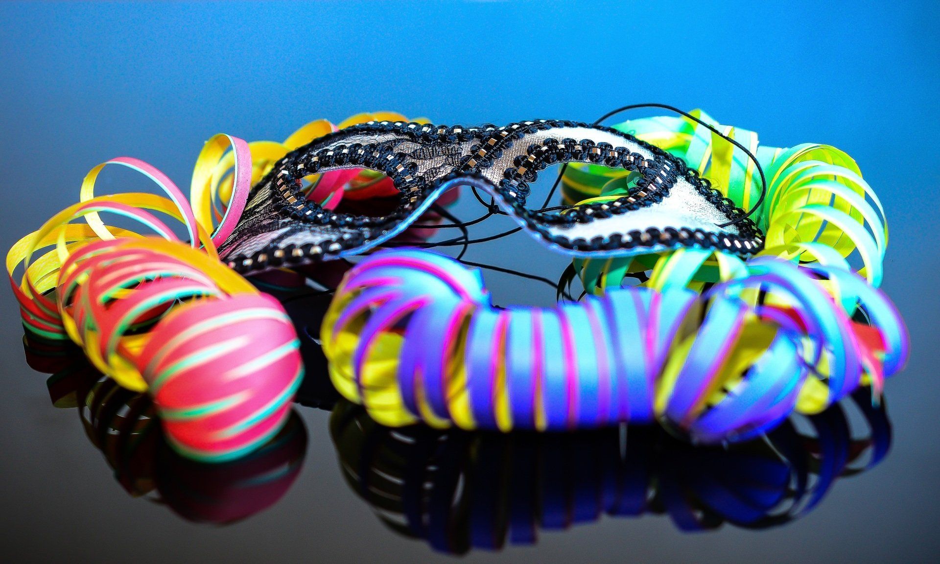 A black masquerade mask resting on a reflective surface, surrounded by colorful, coiled party ribbons against a blue background.