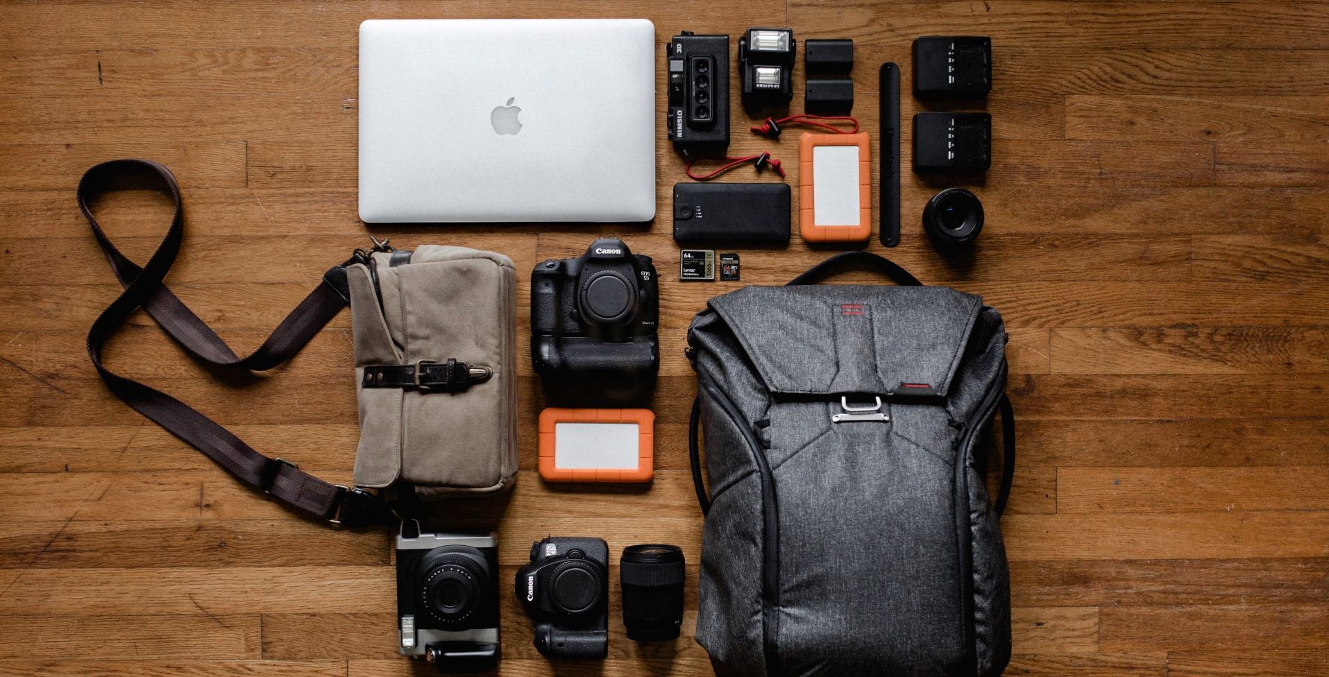 A flat lay of photography gear, including cameras, lenses, a laptop, and a backpack, arranged on a wooden floor.