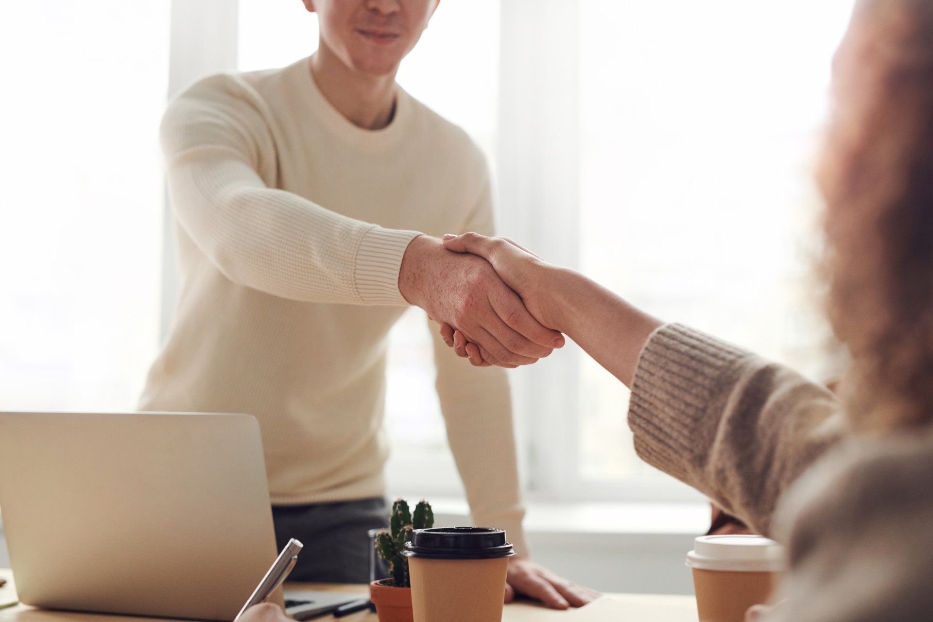 Two people shaking hands across a desk in a bright office, with a laptop and coffee cups on the table.