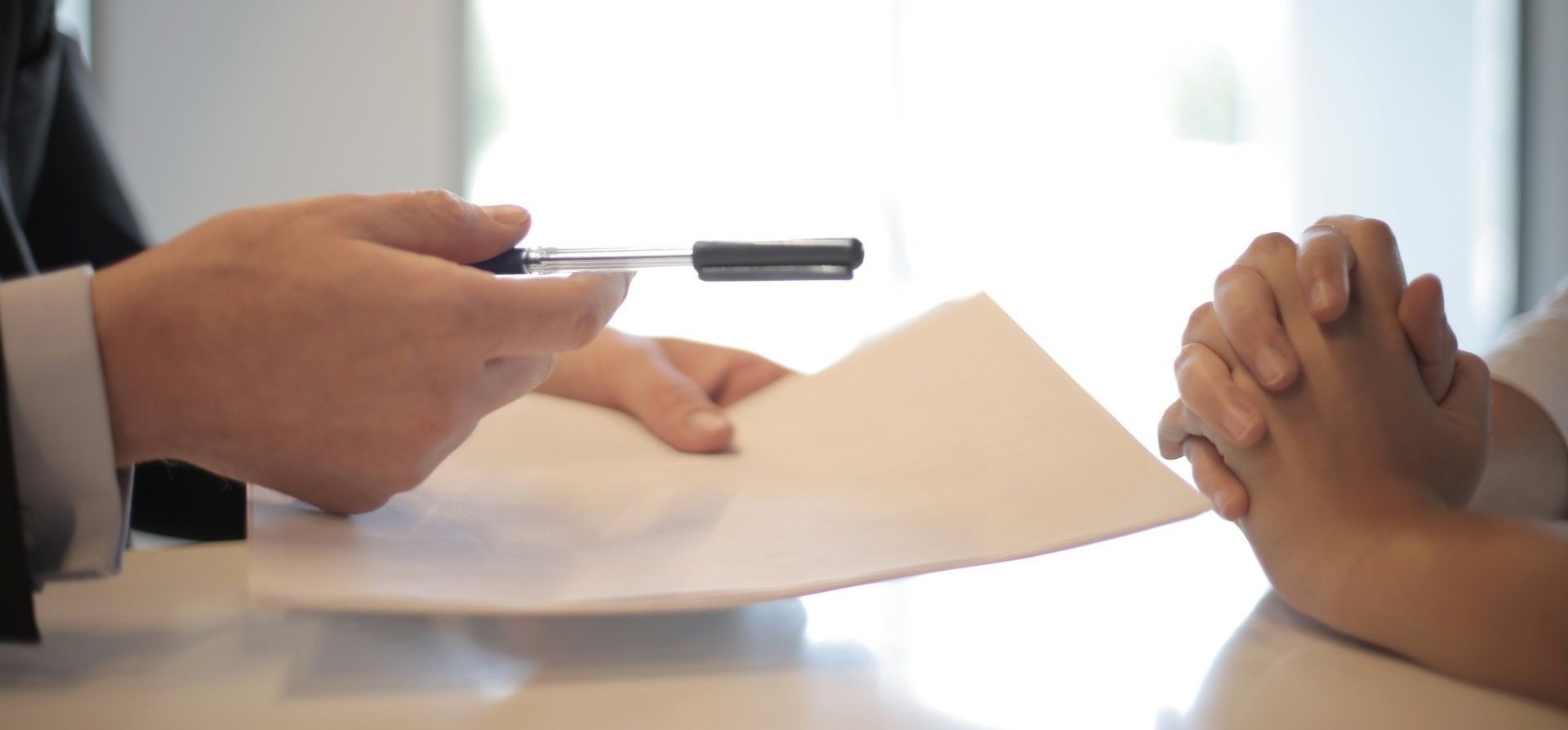A person in a suit holds a pen out over a blank document toward another person sitting across from them at a desk.