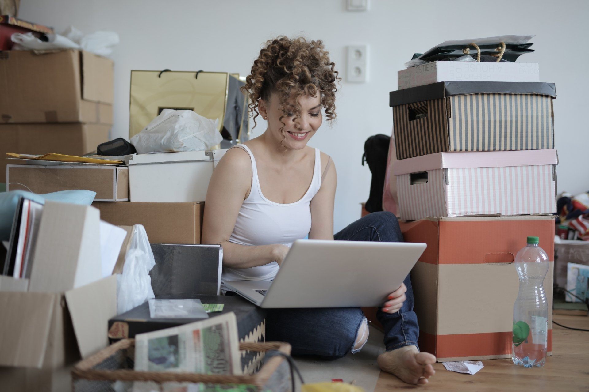 A smiling person sits on the floor surrounded by cardboard boxes while using a laptop.