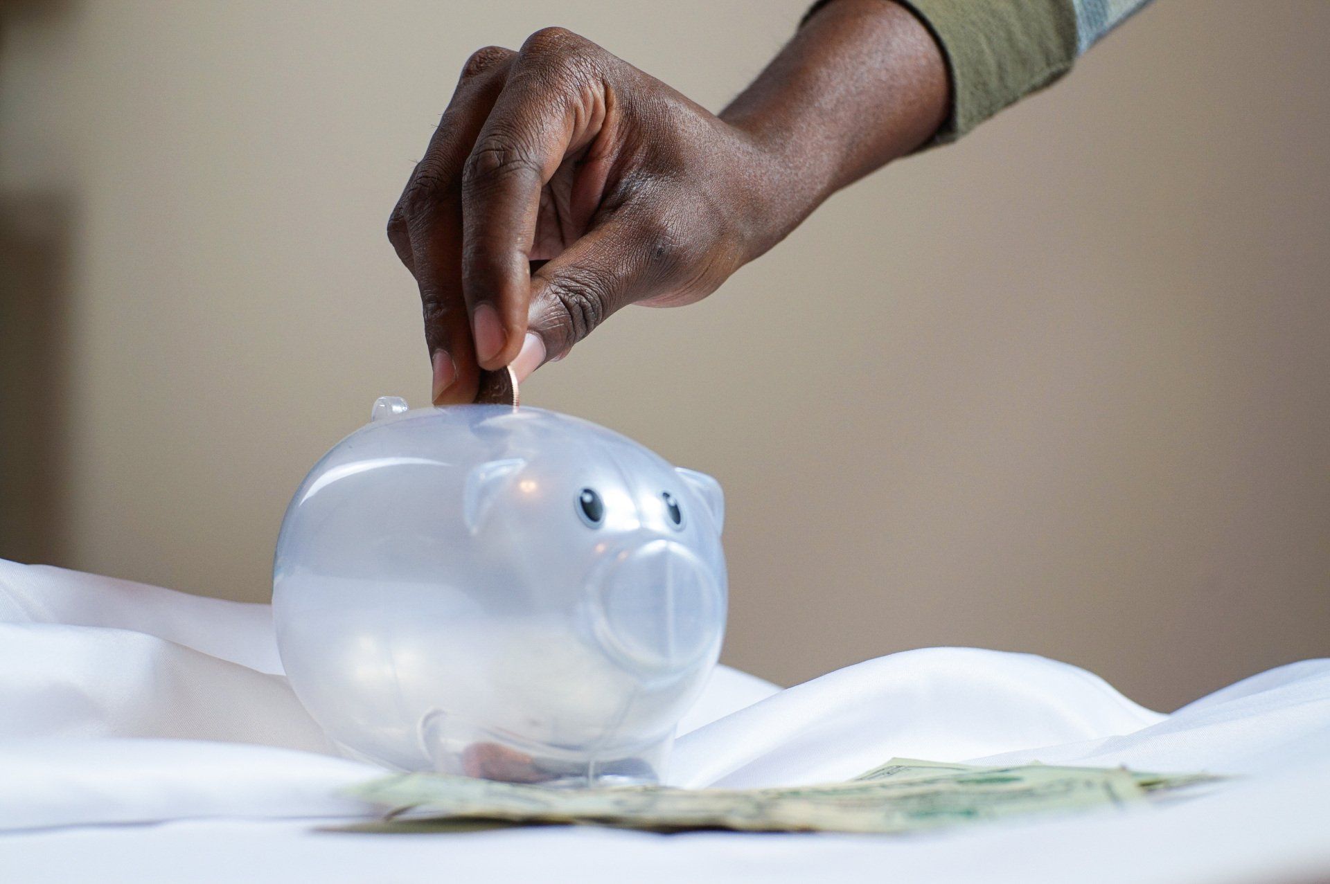 A hand inserts a coin into a translucent piggy bank sitting on a white surface with paper money nearby.