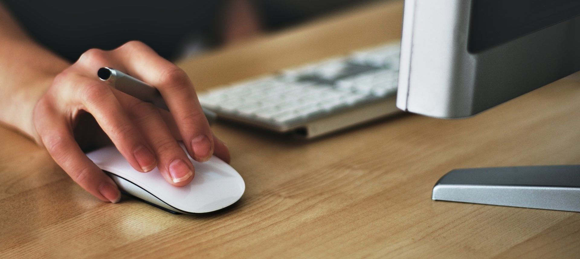A person's hand holding a pen and clicking a white computer mouse on a wooden desk near a keyboard and monitor.