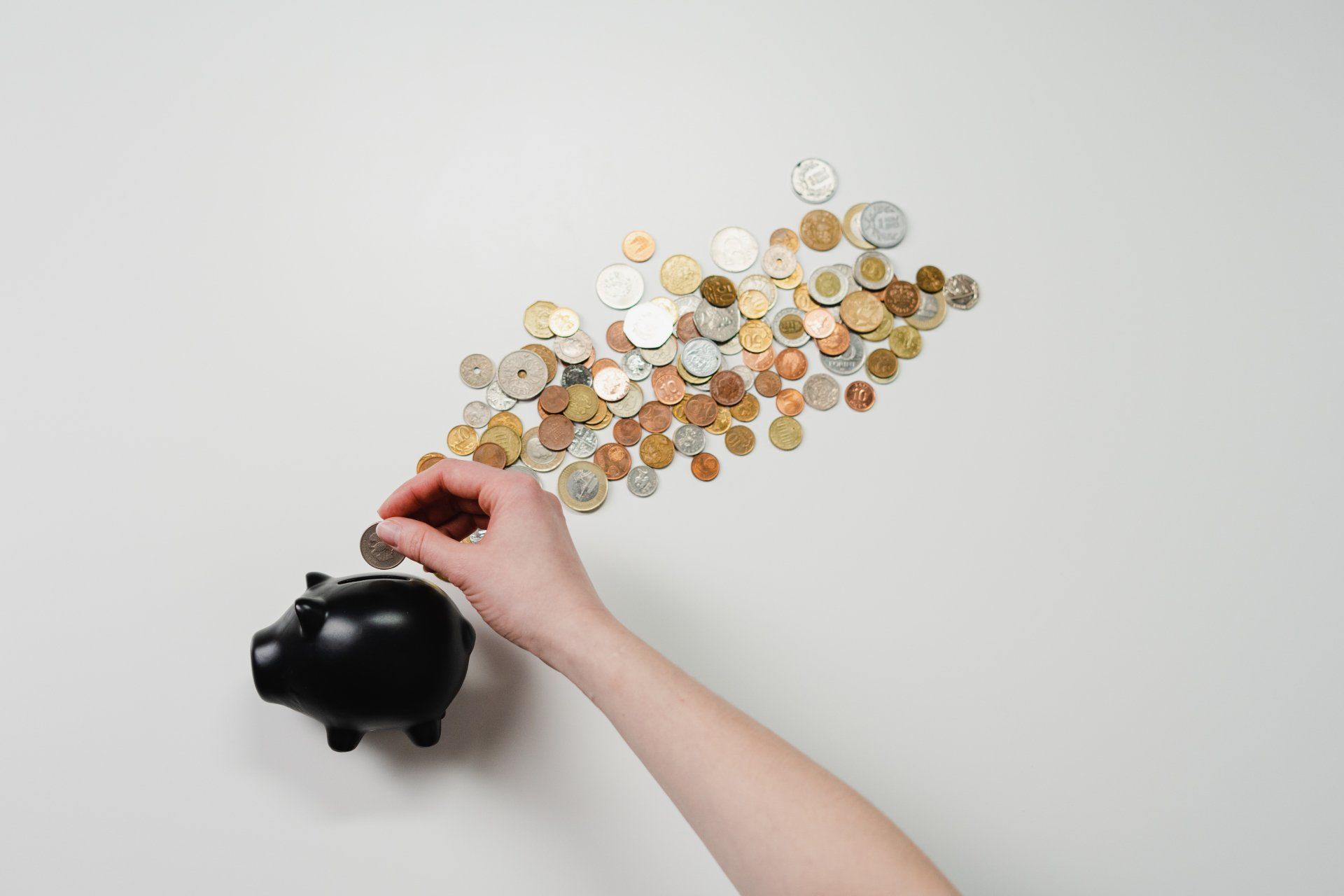 A hand puts a coin into a black piggy bank, with a large collection of scattered coins arranged above it on a white surface.