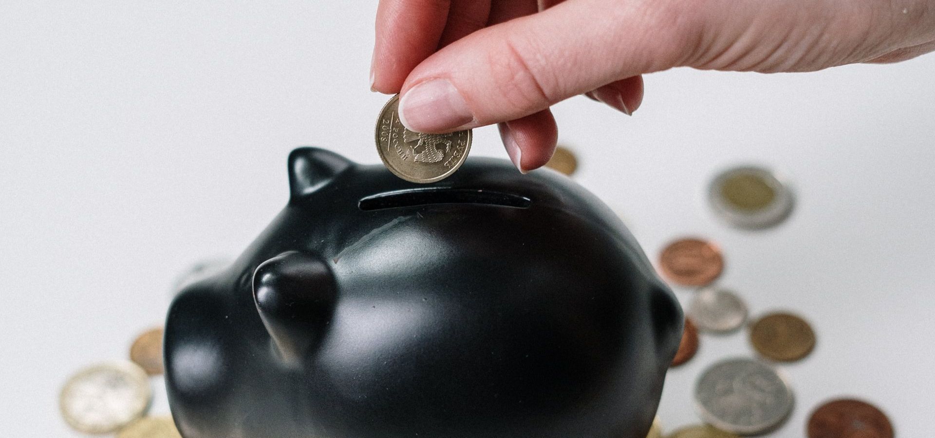 A hand inserts a coin into a black piggy bank surrounded by scattered coins on a white surface.
