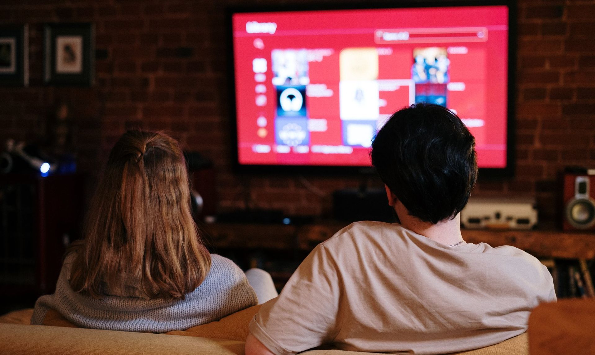 Two people sit on a couch in a dimly lit room, facing a large television screen displaying a media library interface.
