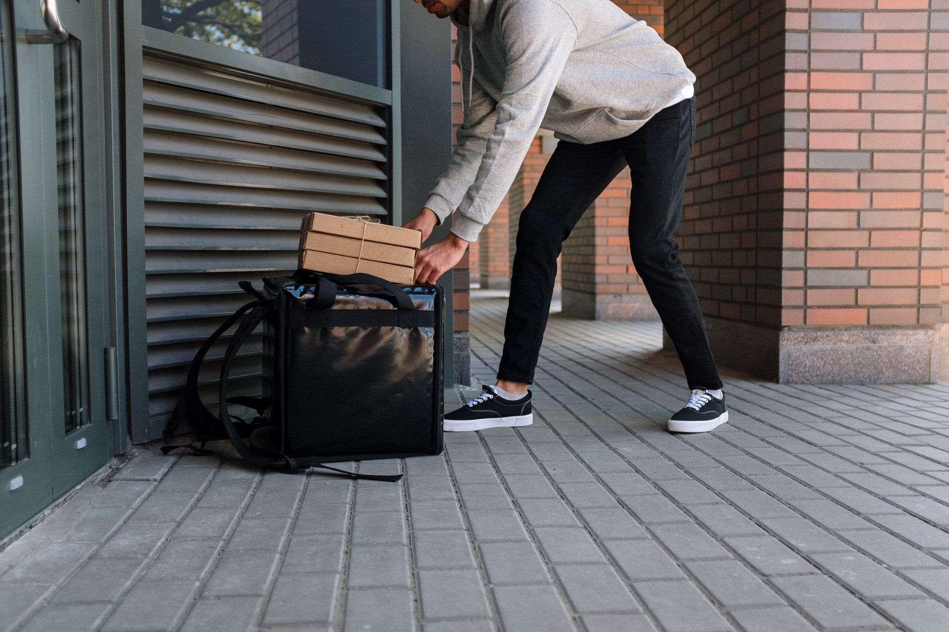 A delivery person in a hoodie and black pants sets a stack of pizza boxes on top of an insulated backpack by a building.