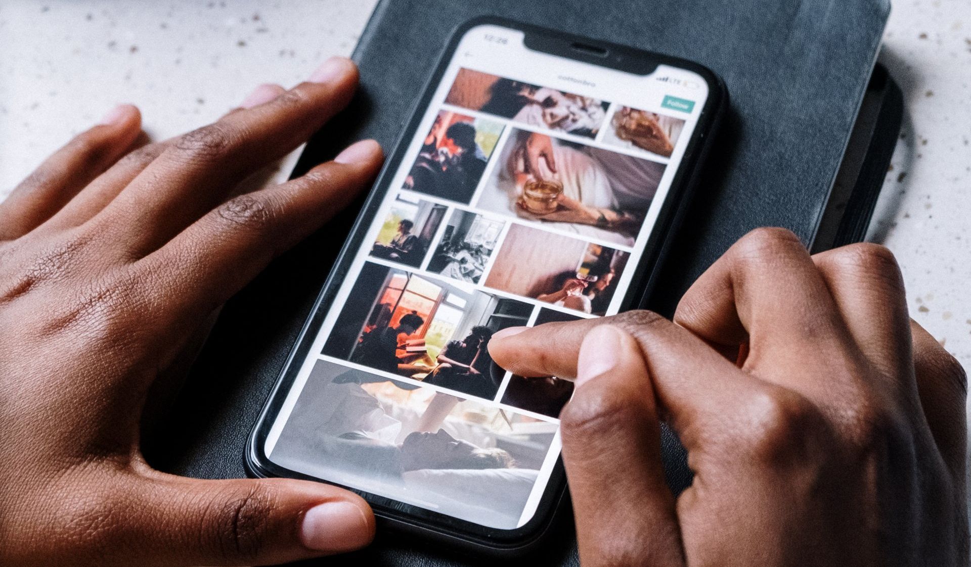 Hands holding a smartphone displaying a grid of photos on a dark, textured background.