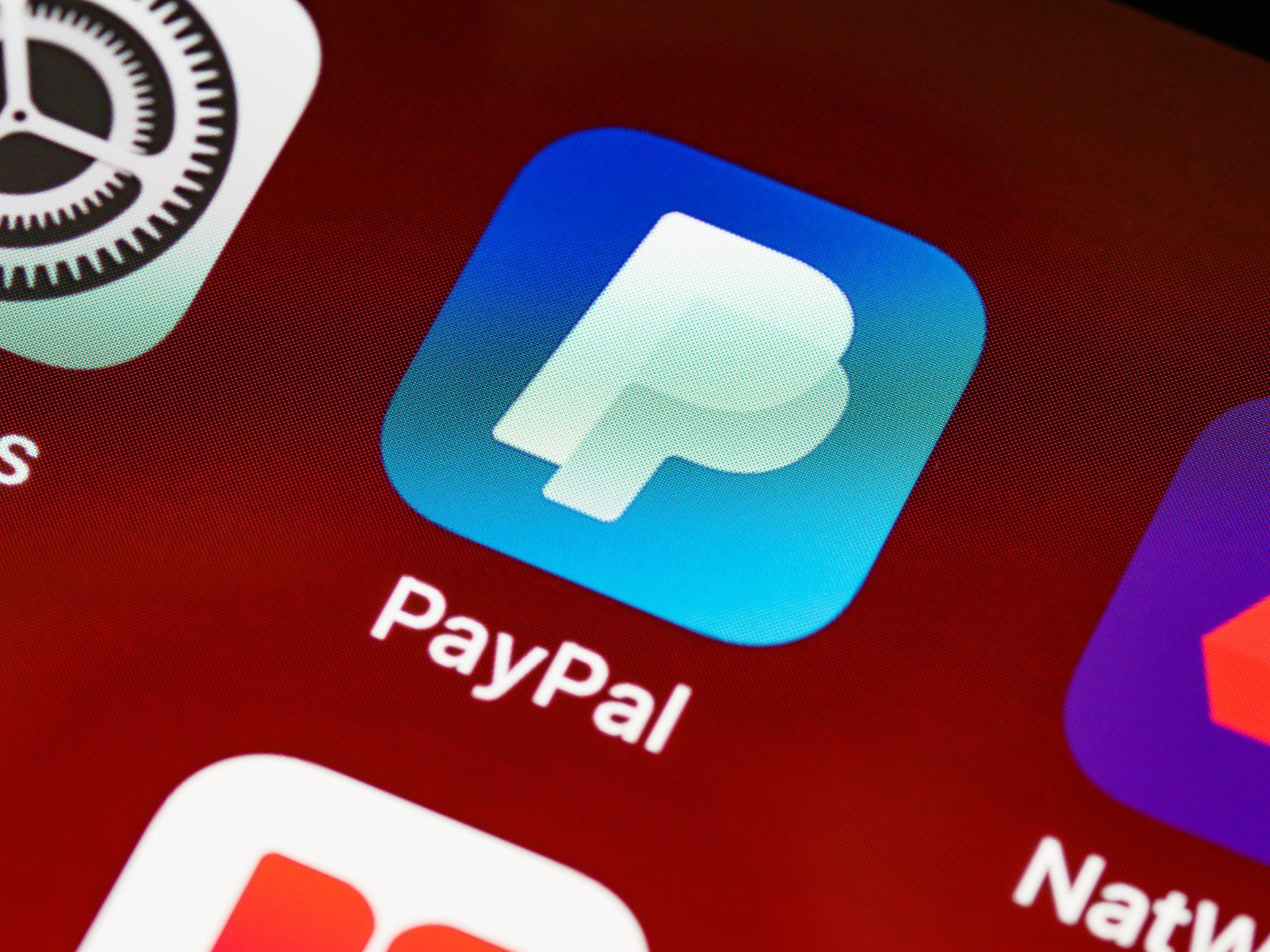 Close-up of a blue PayPal app icon on a smartphone home screen, surrounded by other app icons on a red background.