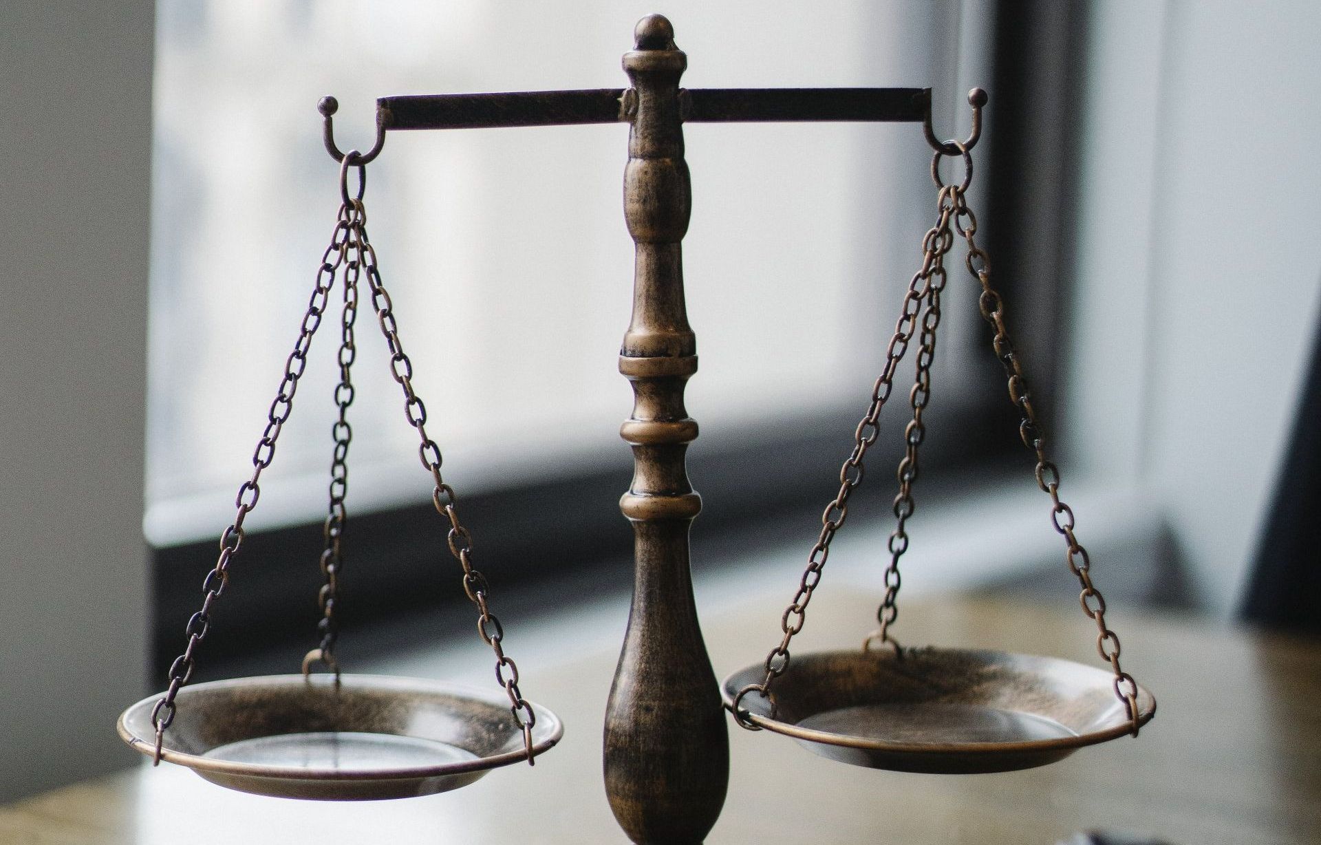 A vintage metal balance scale stands on a wooden surface, with two empty pans suspended by chains against a blurry window.