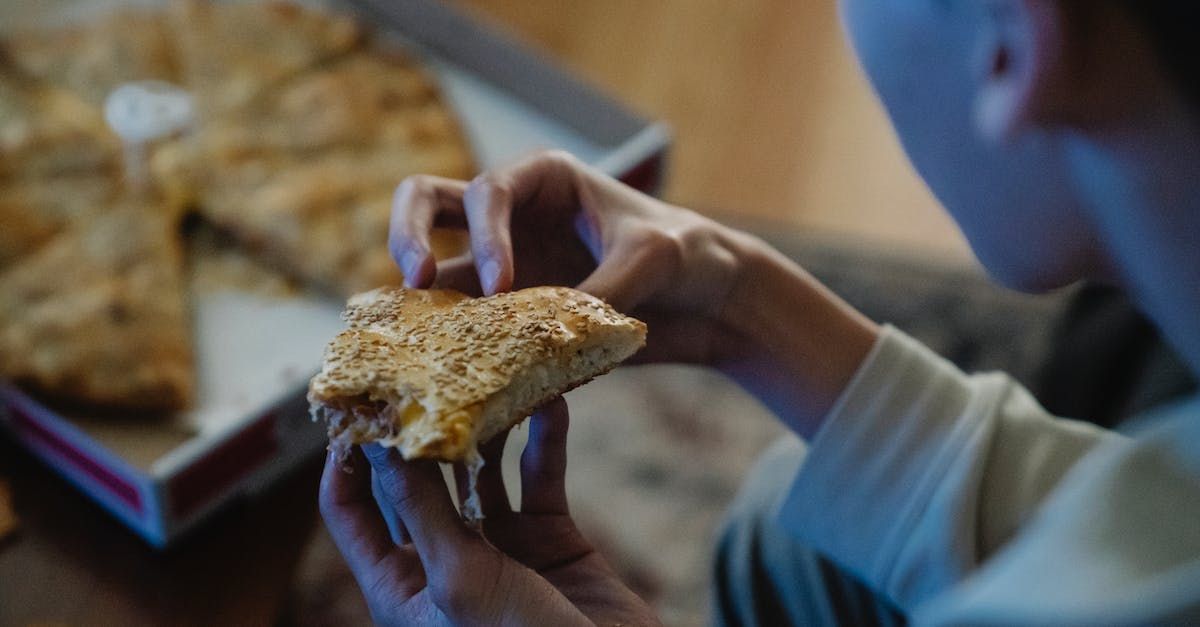 A person holds a slice of pizza in their hands, with a pizza box in the blurred background.