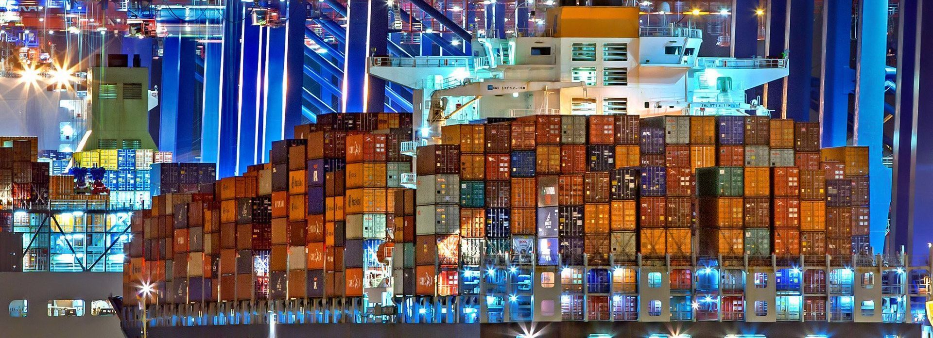 A massive cargo ship heavily loaded with stacked shipping containers at a brightly lit port at night.