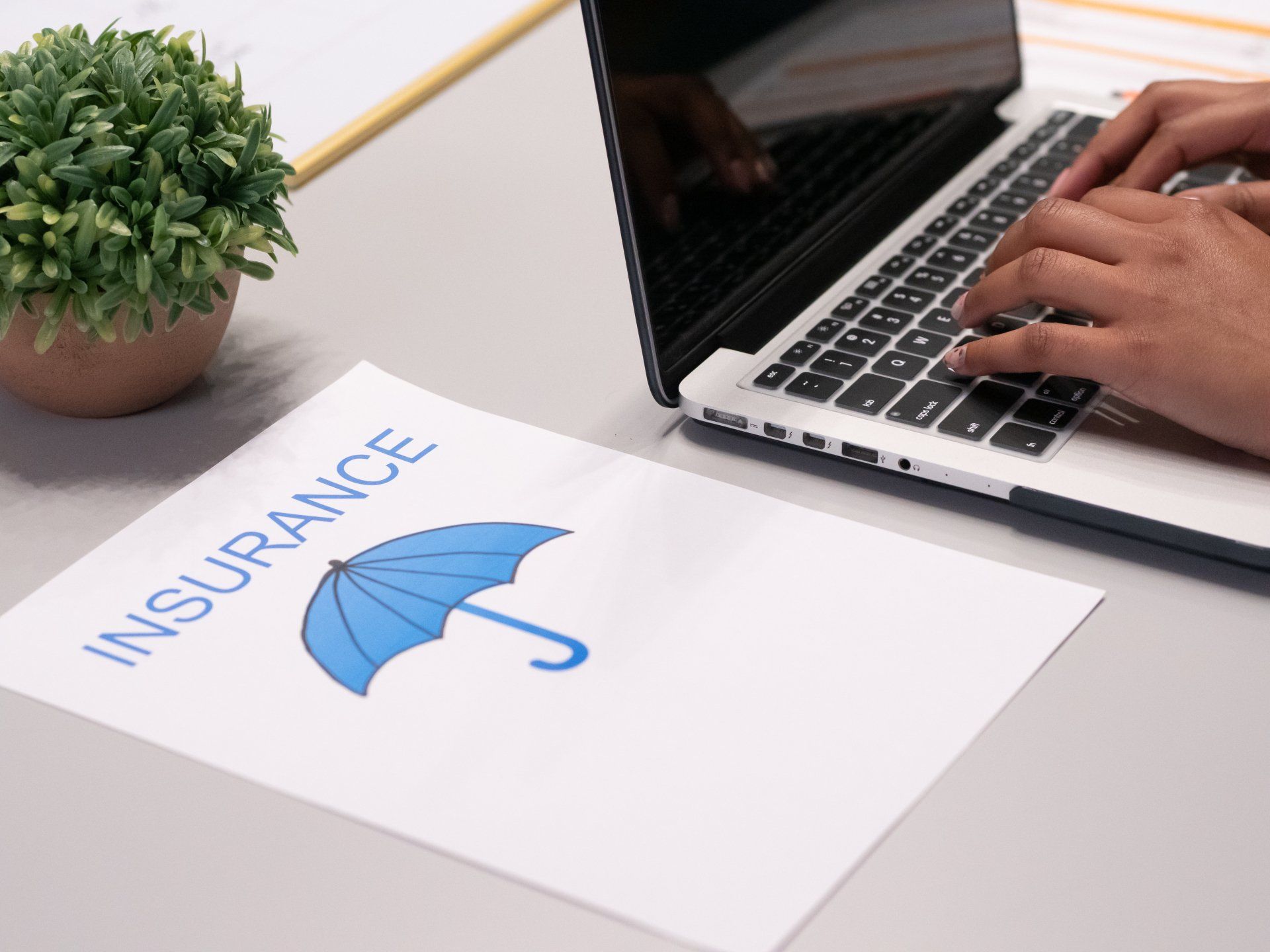A person types on a laptop next to a small potted plant and a paper with the word 
