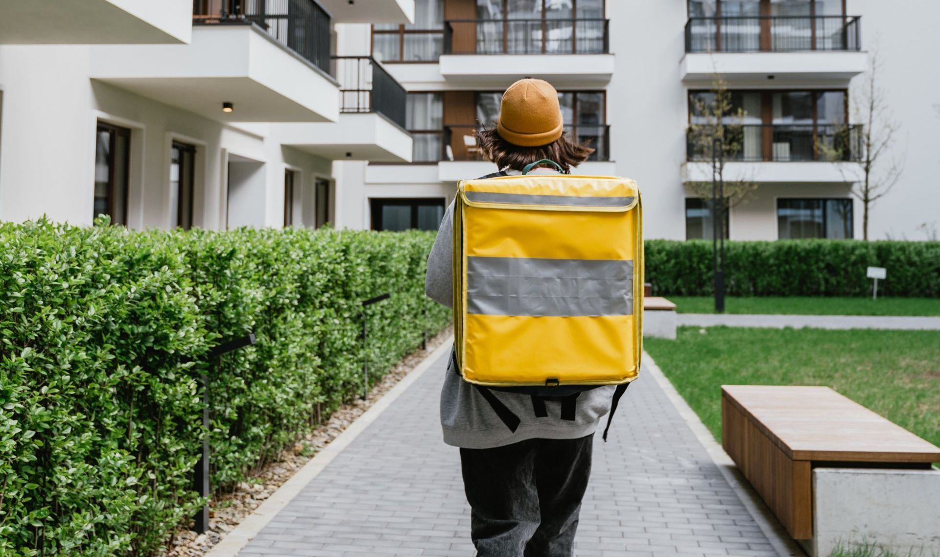 A person with a yellow delivery backpack walks along a stone path toward a modern apartment building.