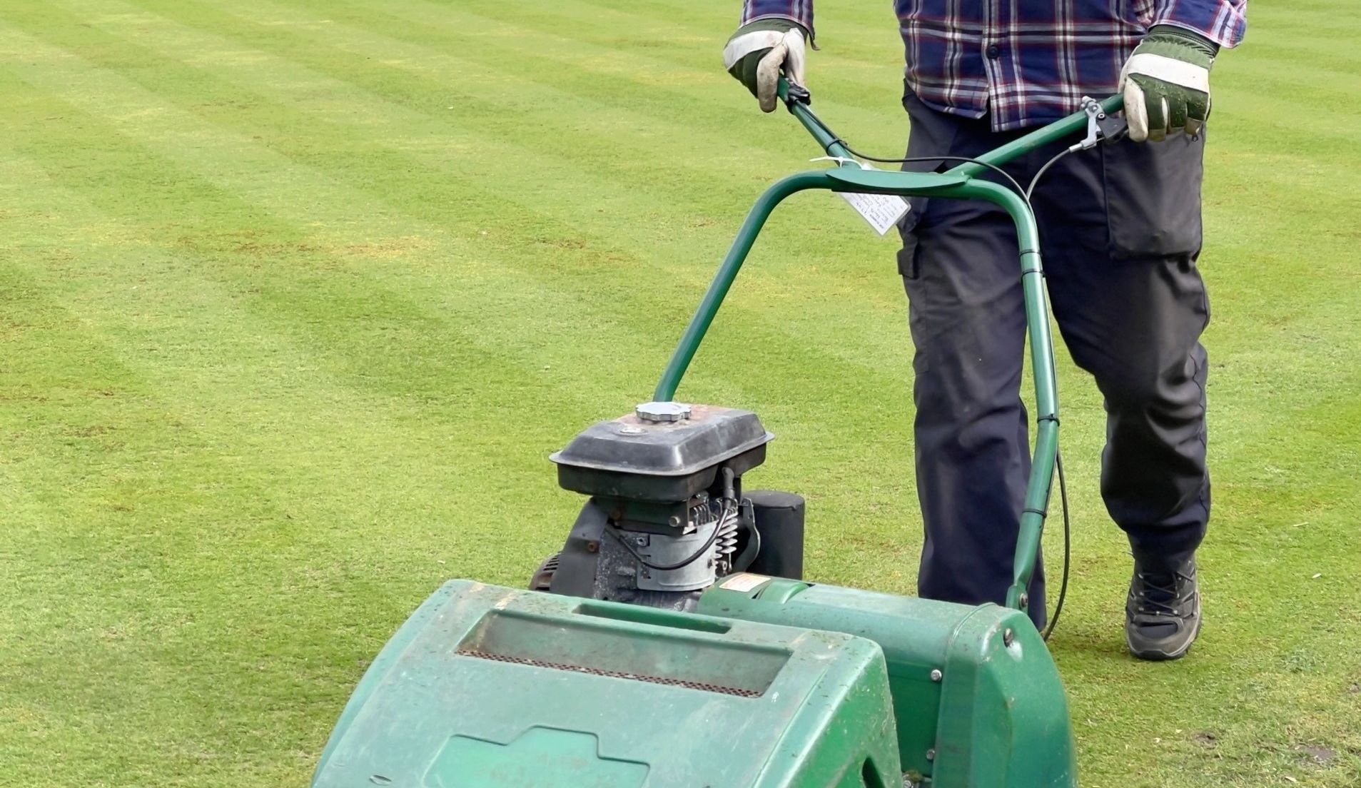 A person pushes a green reel mower across a manicured lawn, leaving stripes in the grass.