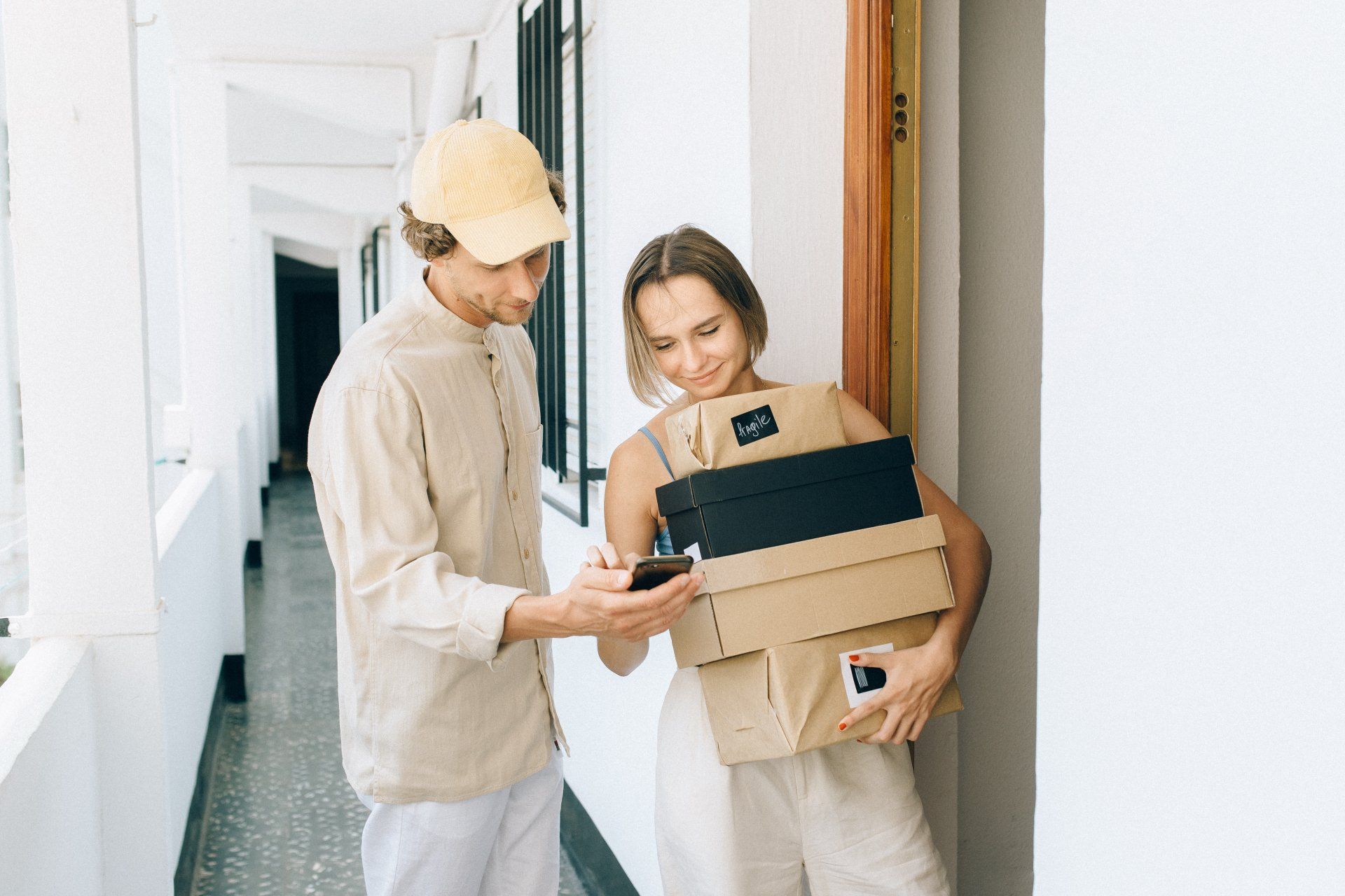 A person in a tan cap shows a smartphone to a person holding a stack of cardboard boxes in a bright, modern hallway.