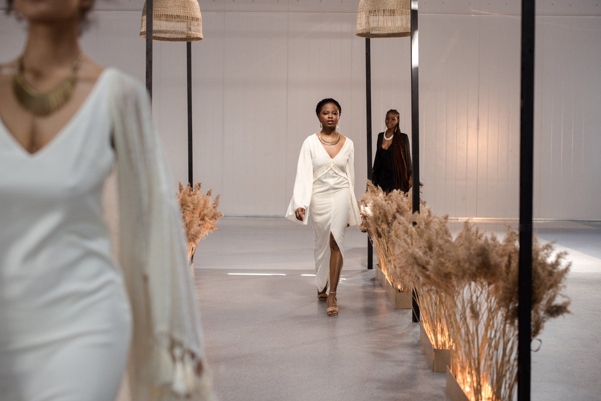 Models walk a minimalist runway lined with dried wheat grass under hanging basket lights in a bright, modern hall.
