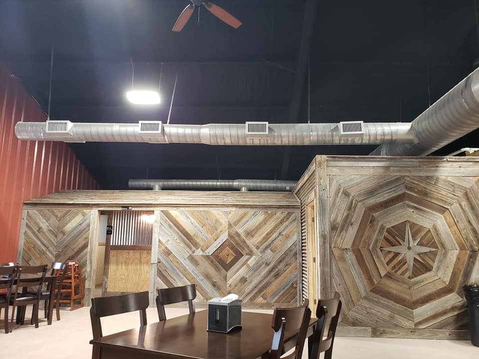 A rustic indoor dining area features geometric reclaimed wood walls and exposed ventilation ducts on a dark ceiling.