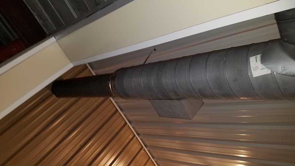 A metallic industrial duct section connected to a dark pipe, suspended near the ceiling of a corrugated metal structure.