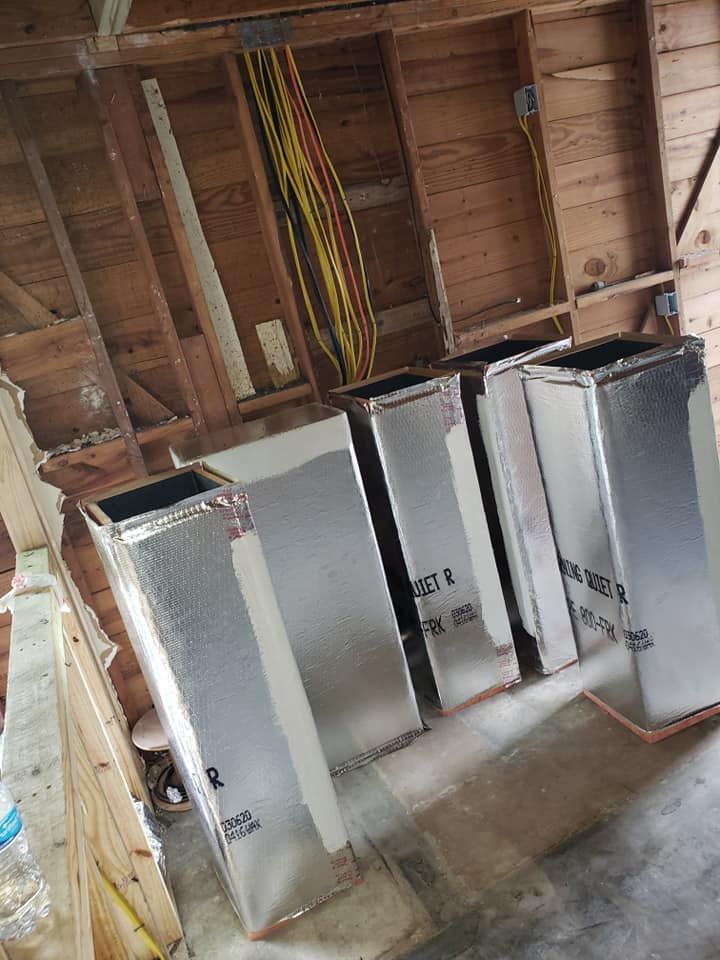 Four rectangular air duct segments wrapped in silver reflective insulation standing on a wooden floor in a framed room.