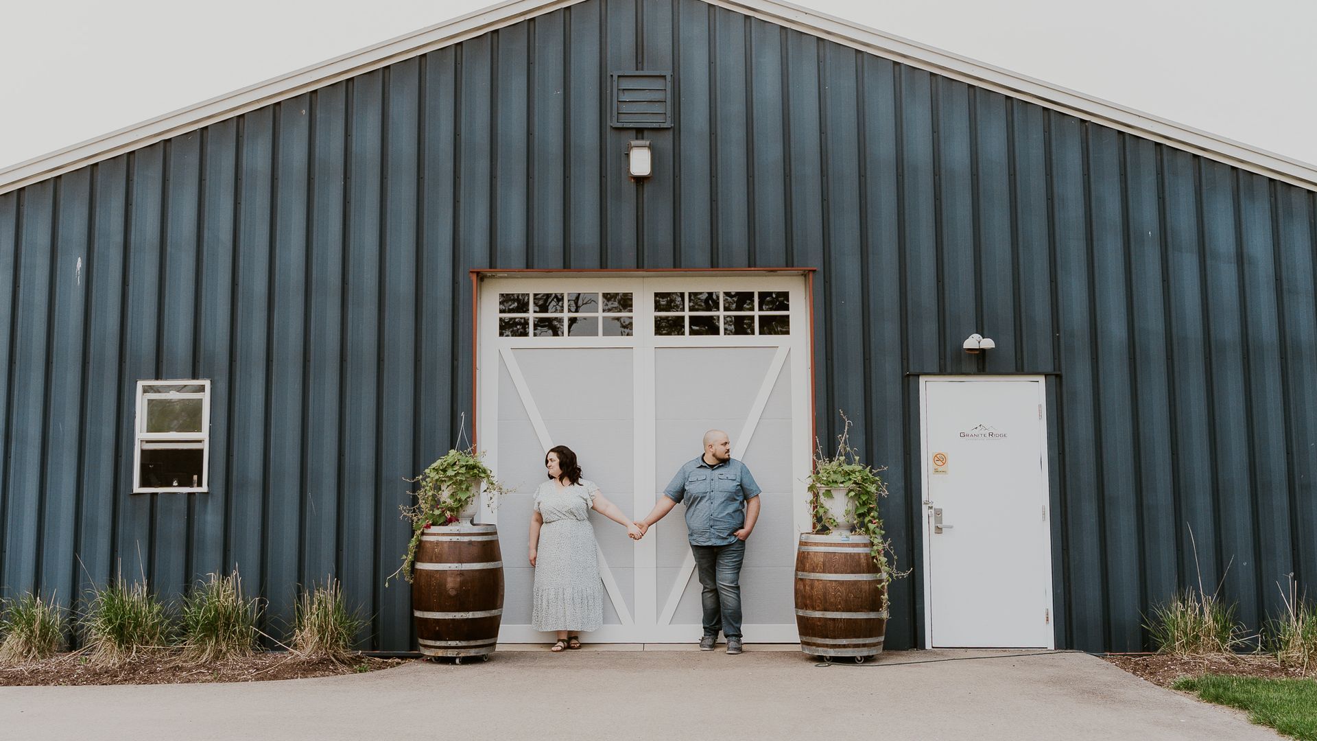 A man and a woman are standing in front of a barn holding hands.