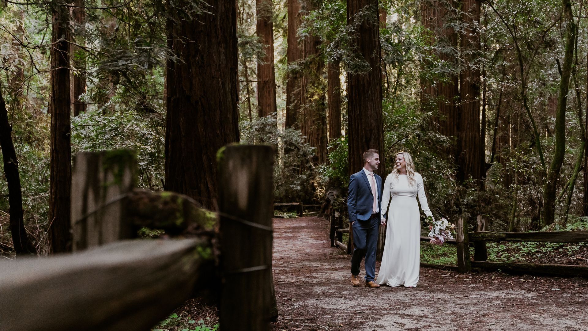 A bride and groom are walking down a path in the woods.