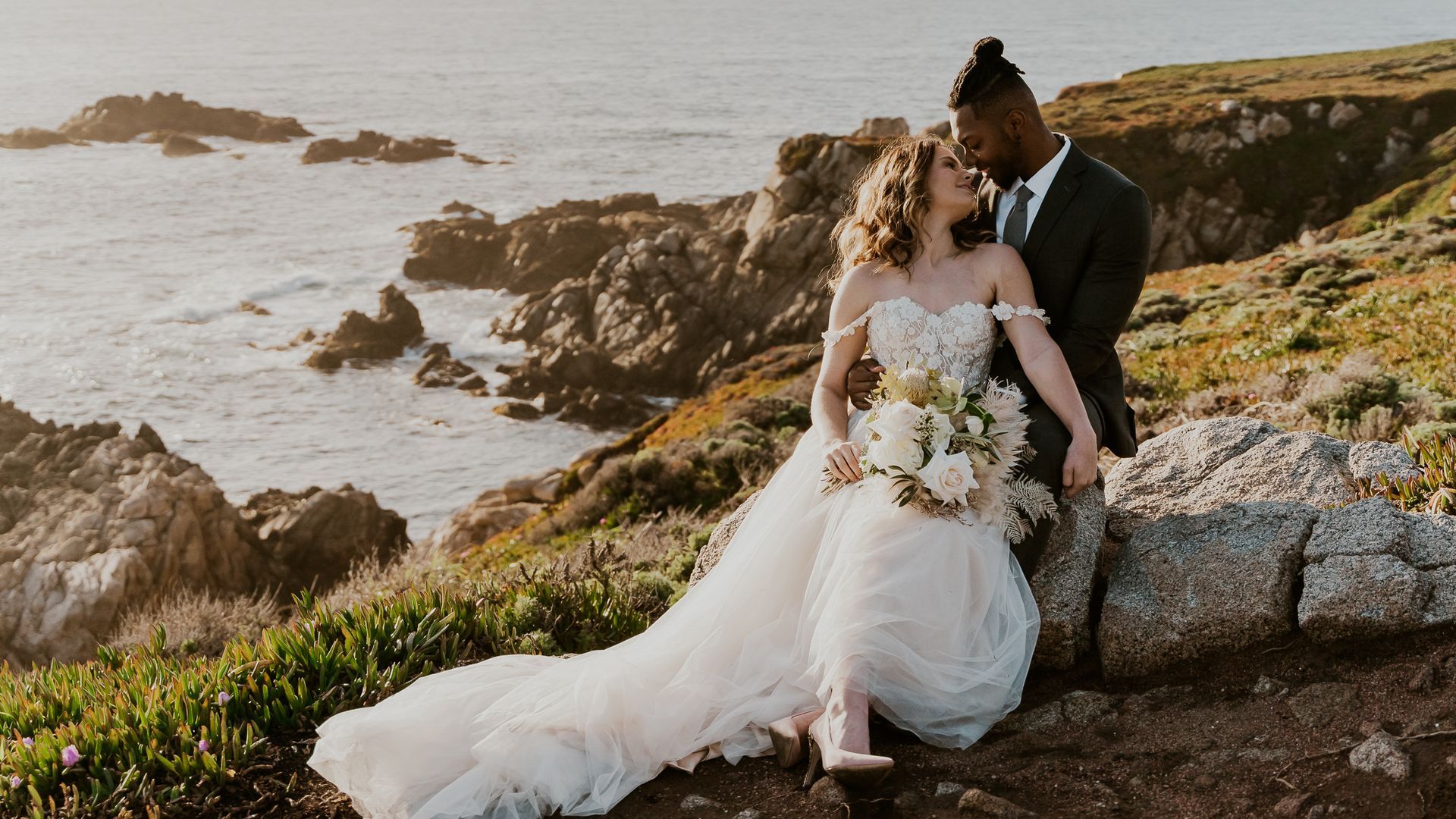 A bride and groom are sitting on a rock near the ocean.