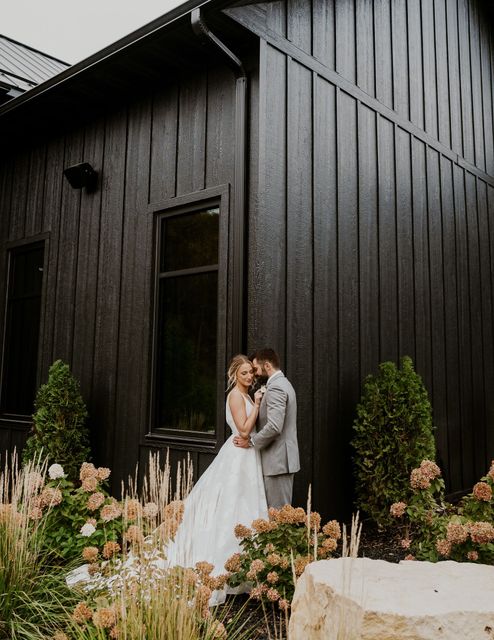 Bride and groom embracing outside a black barn, surrounded by white and pink flowers.