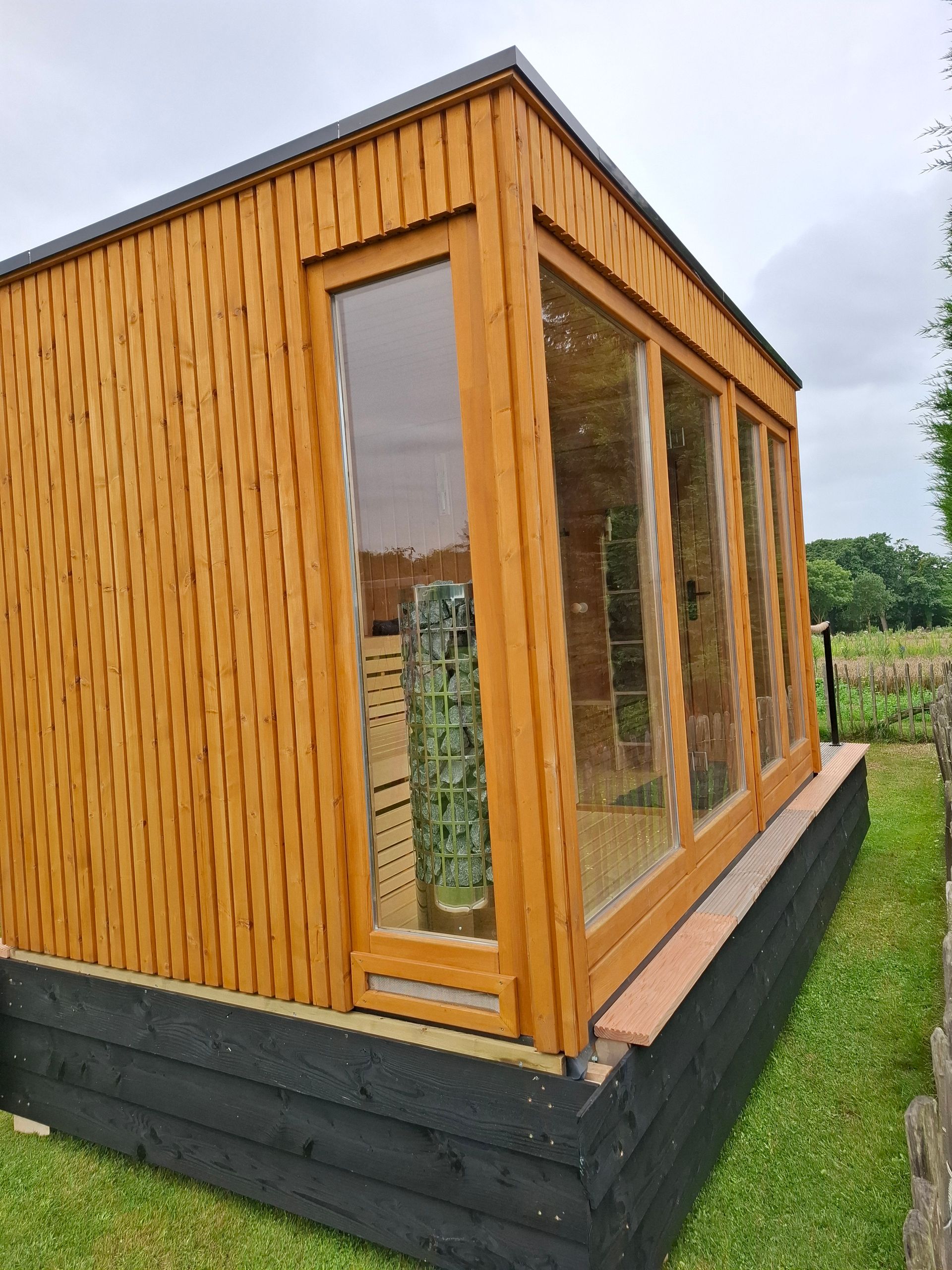 Houten tuinhuis met grote ramen, geplaatst op een verhoogd platform, omgeven door gras.