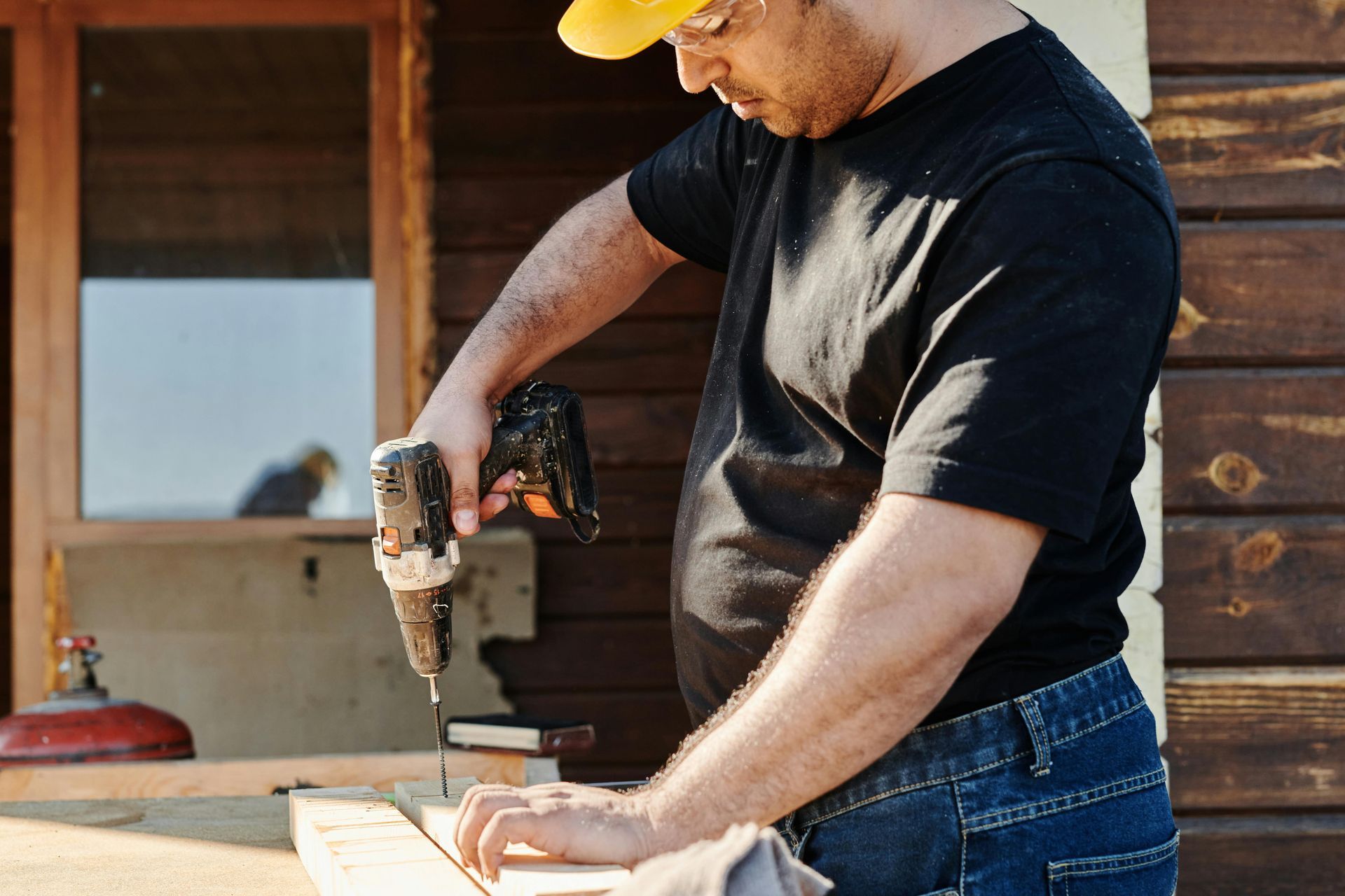 Man in yellow hard hat drills wood on a table outdoors near a wooden structure.