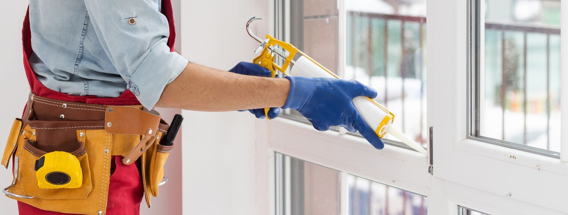 Person applying caulk to a window frame using a caulking gun while wearing gloves.