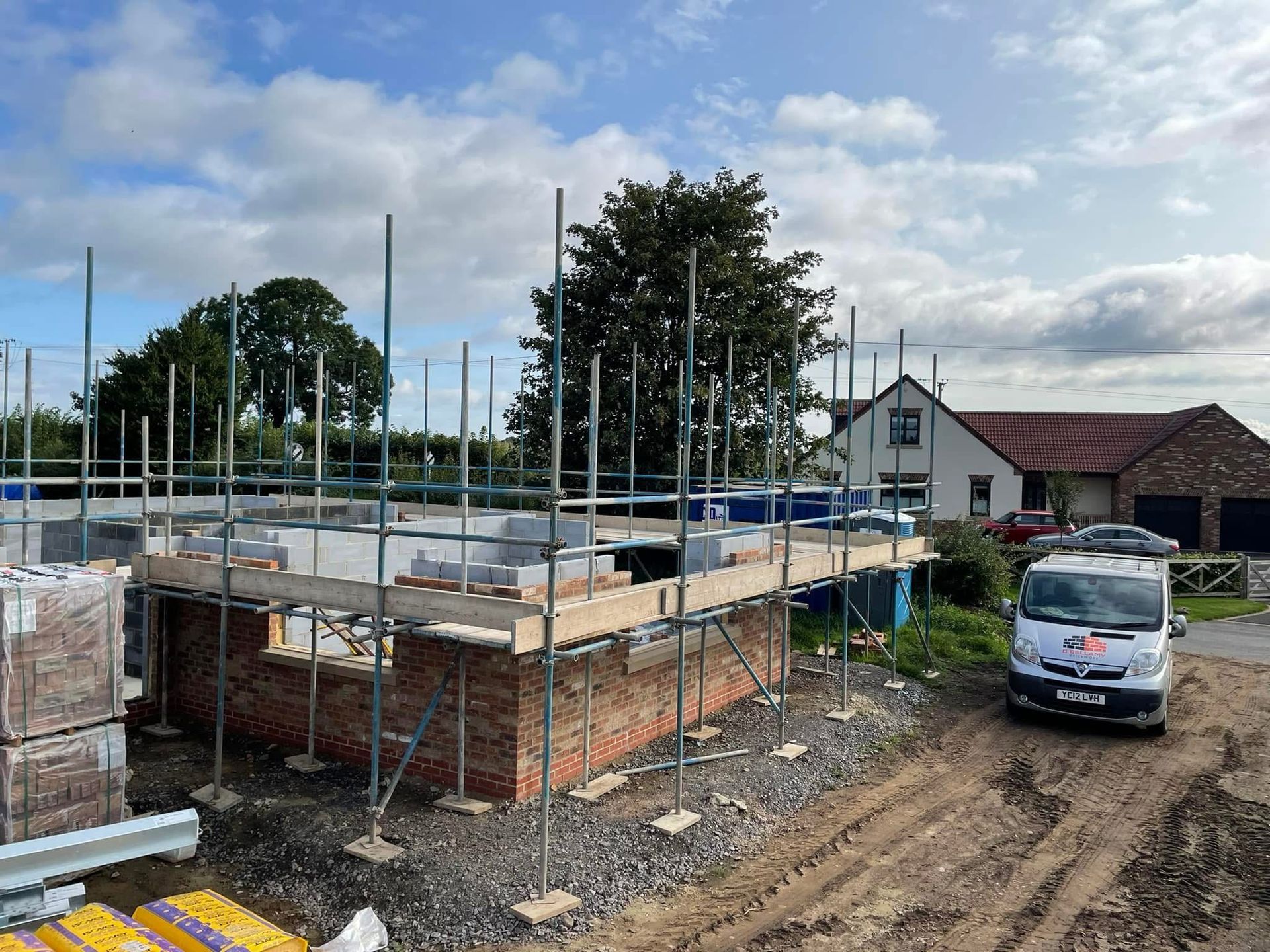 Construction site: Brick building with scaffolding, materials, and a work van on a dirt lot under a cloudy sky.
