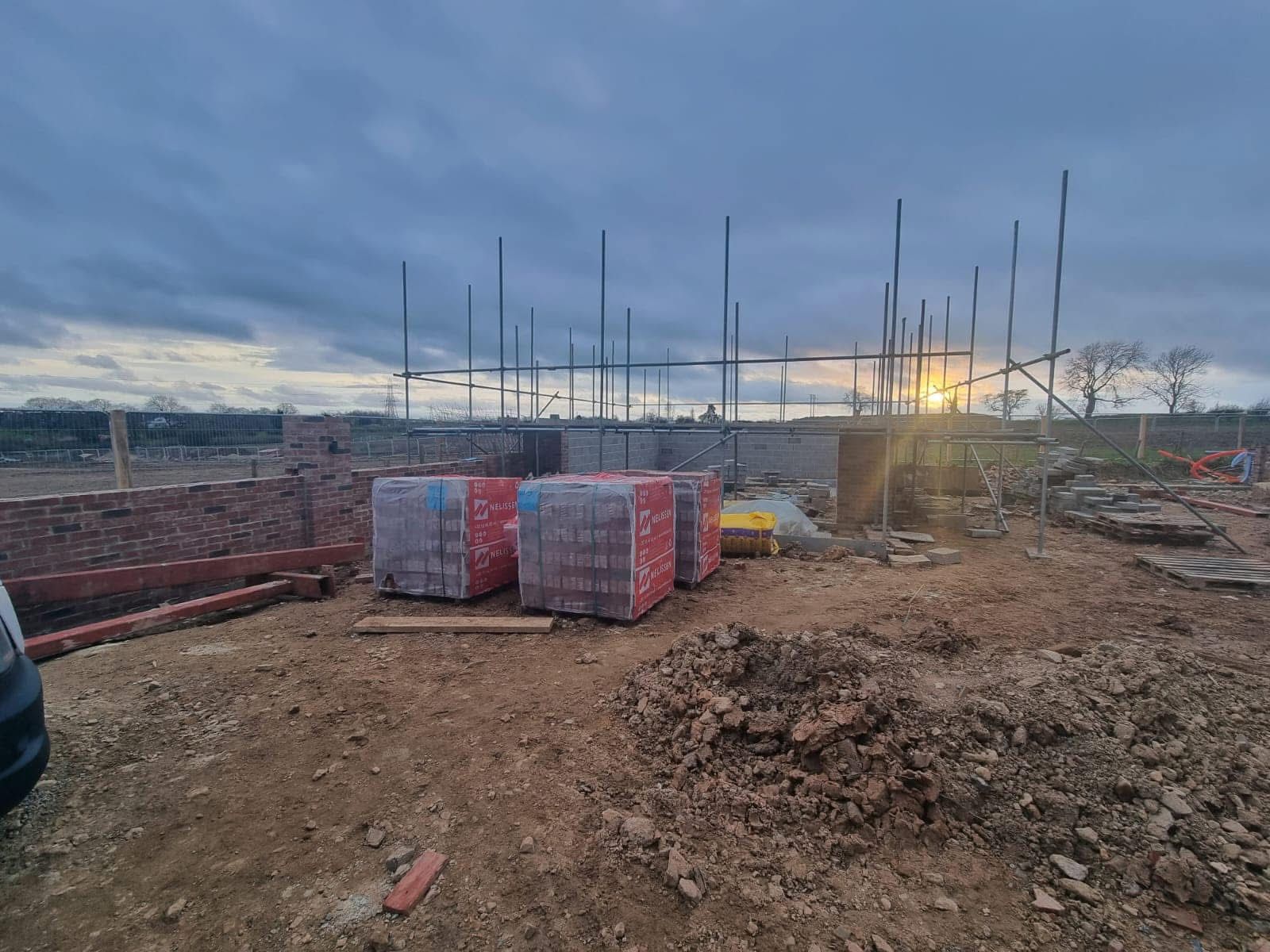 Construction site with brick walls, scaffolding, and stacks of bricks under a cloudy sky at sunset.