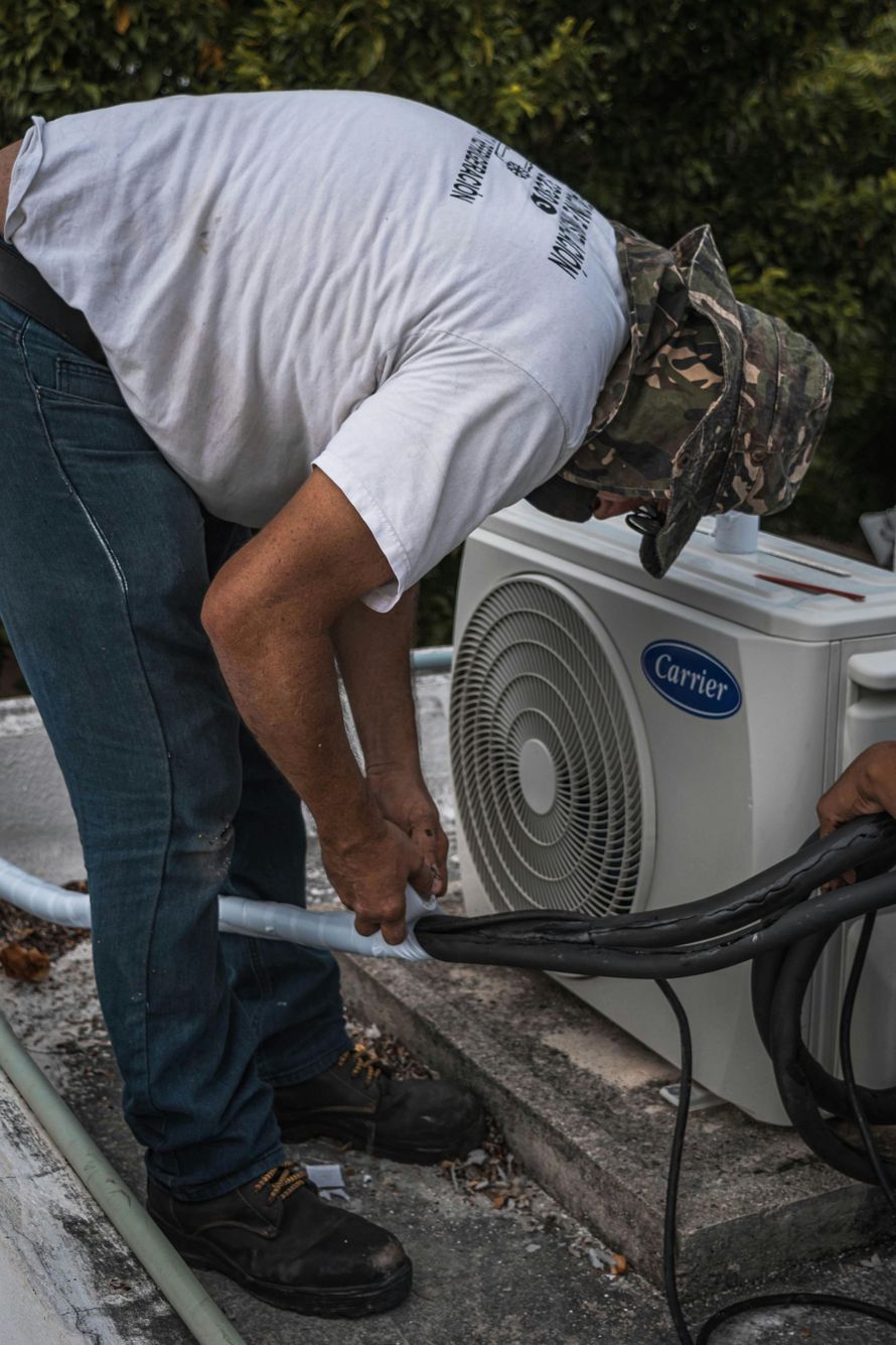 A technician wearing a camouflage hat works on a Carrier outdoor air conditioning unit on a rooftop.
