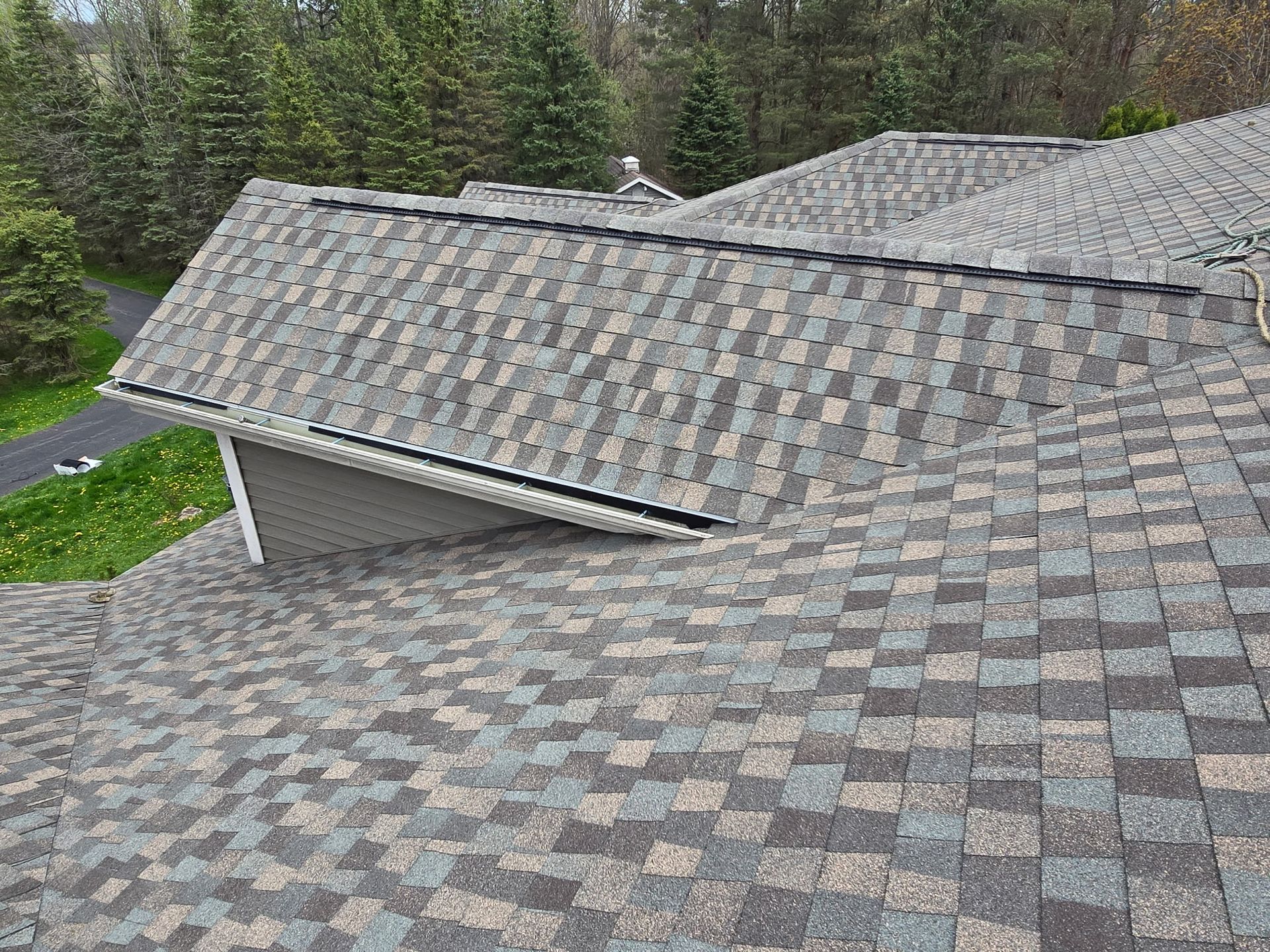 An overhead view of a gray-toned, multi-textured shingled roof with several gables, set against a background of trees.