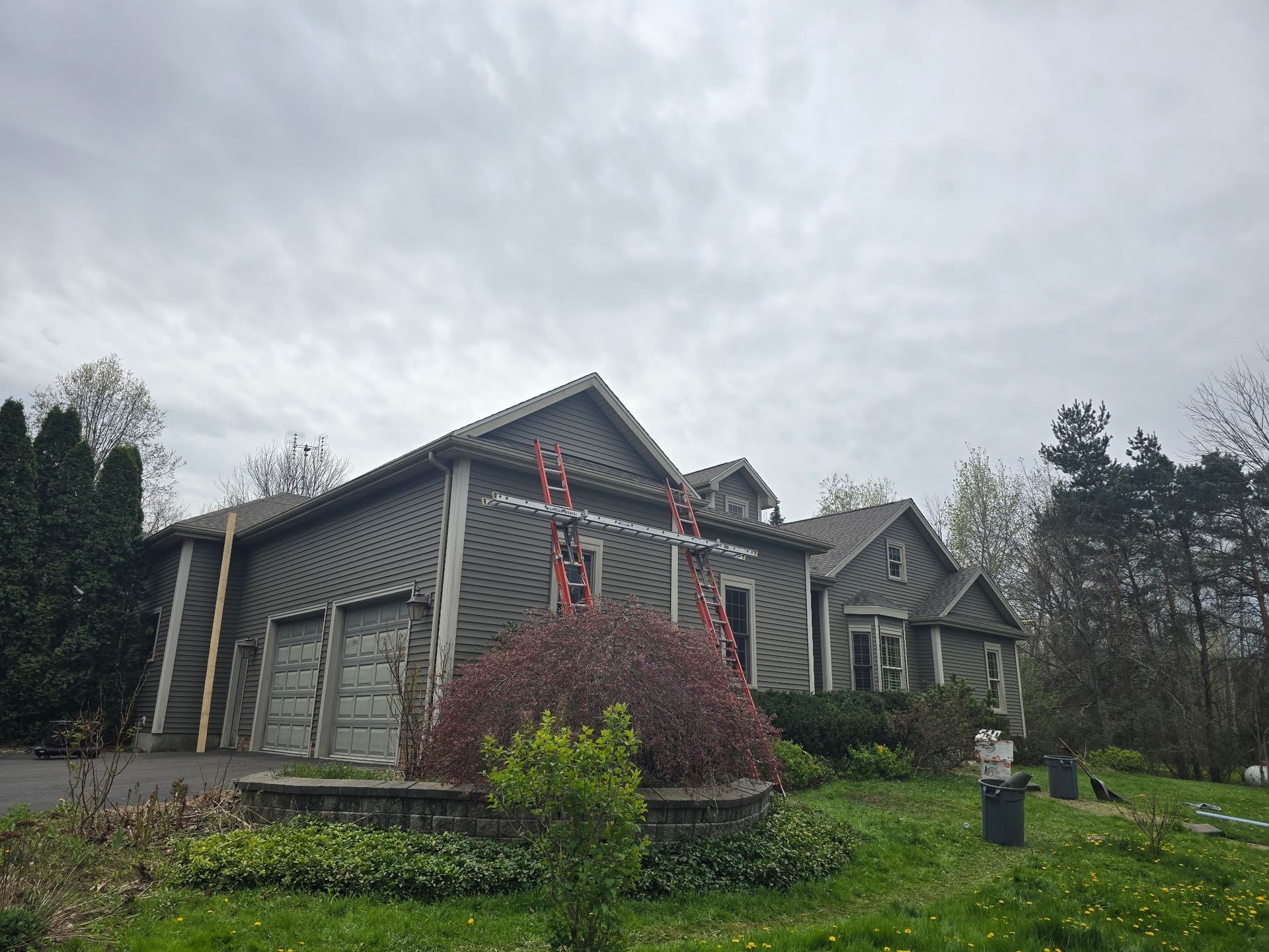 A two-story grey-sided house with a garage, surrounded by trees and overgrown landscaping under a cloudy sky.