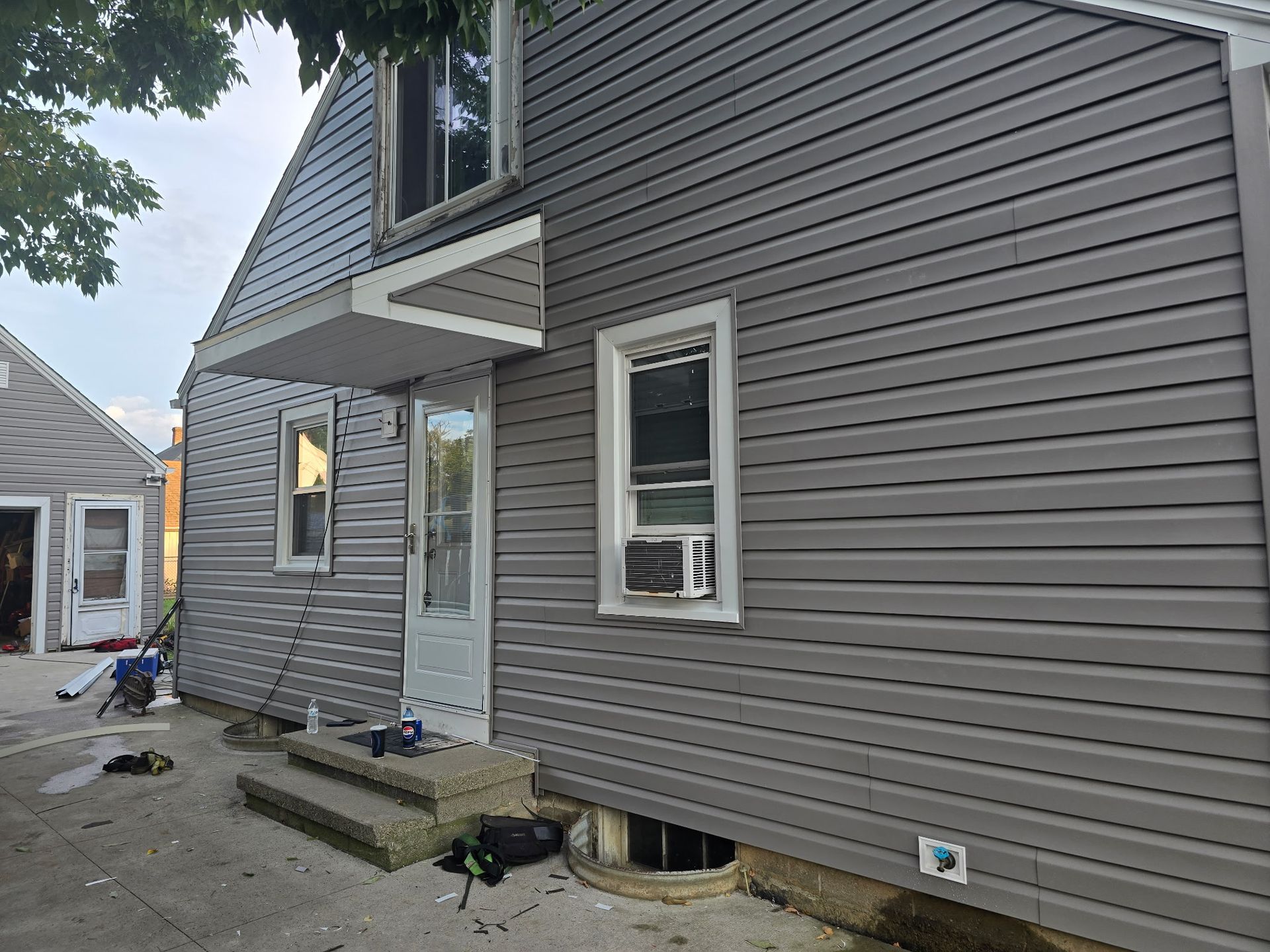 Side view of a grey-sided house with a white door, small awning, windows, and concrete steps.