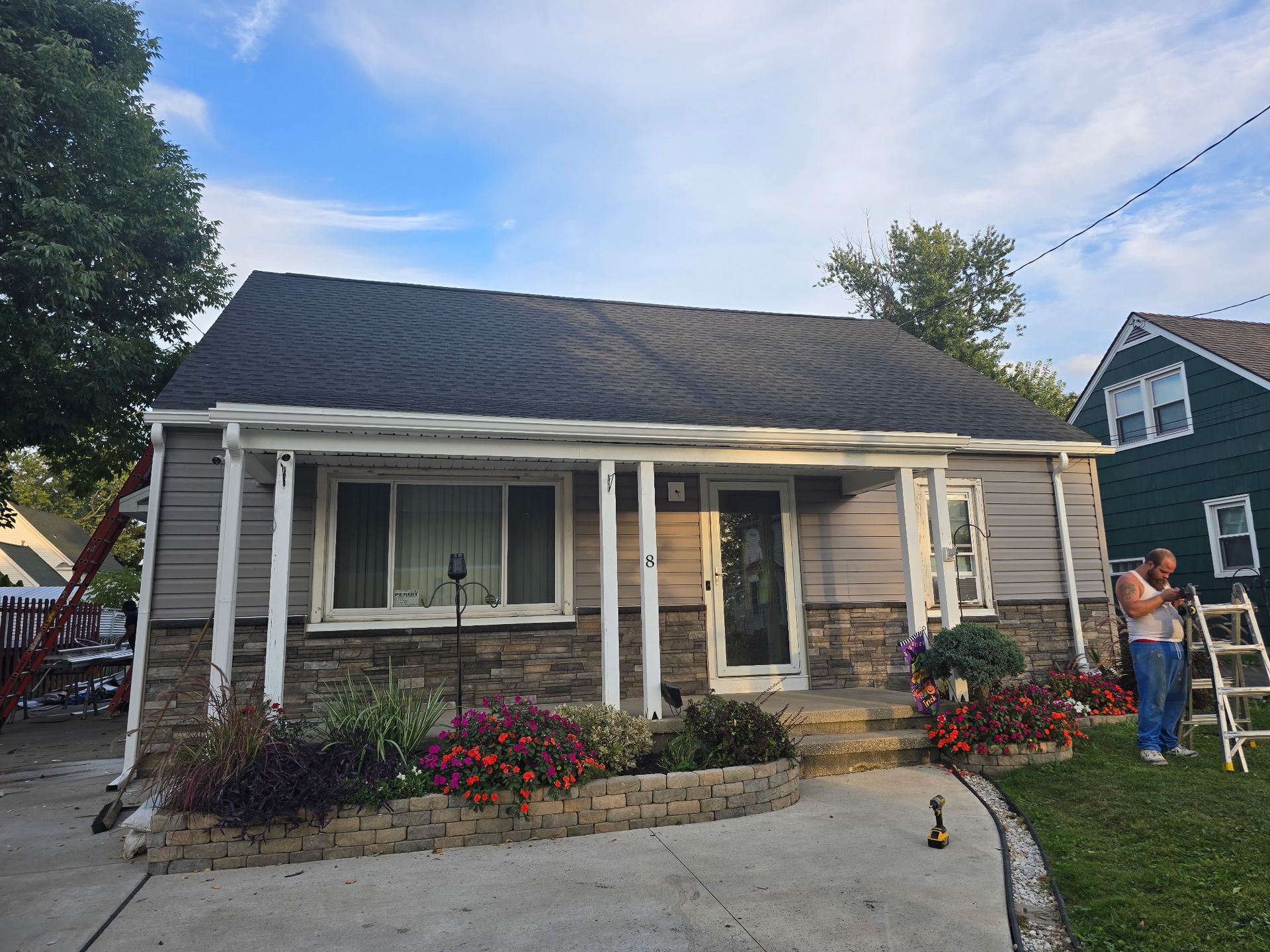 A single-story house with gray siding and stone accents, featuring a front porch with a person working on a ladder.