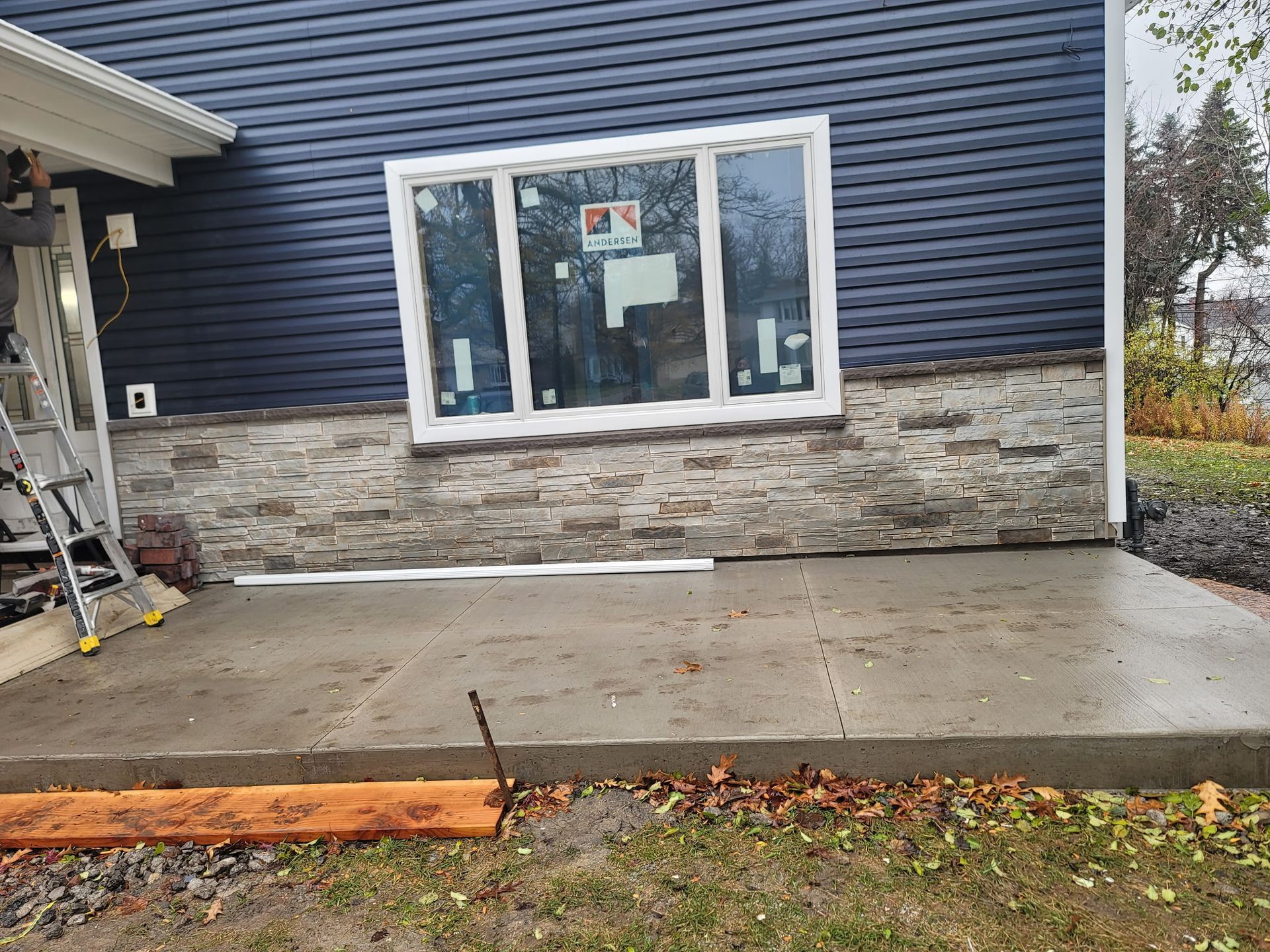 A freshly poured concrete patio in front of a blue-sided house with a stone veneer foundation and a white-framed window.
