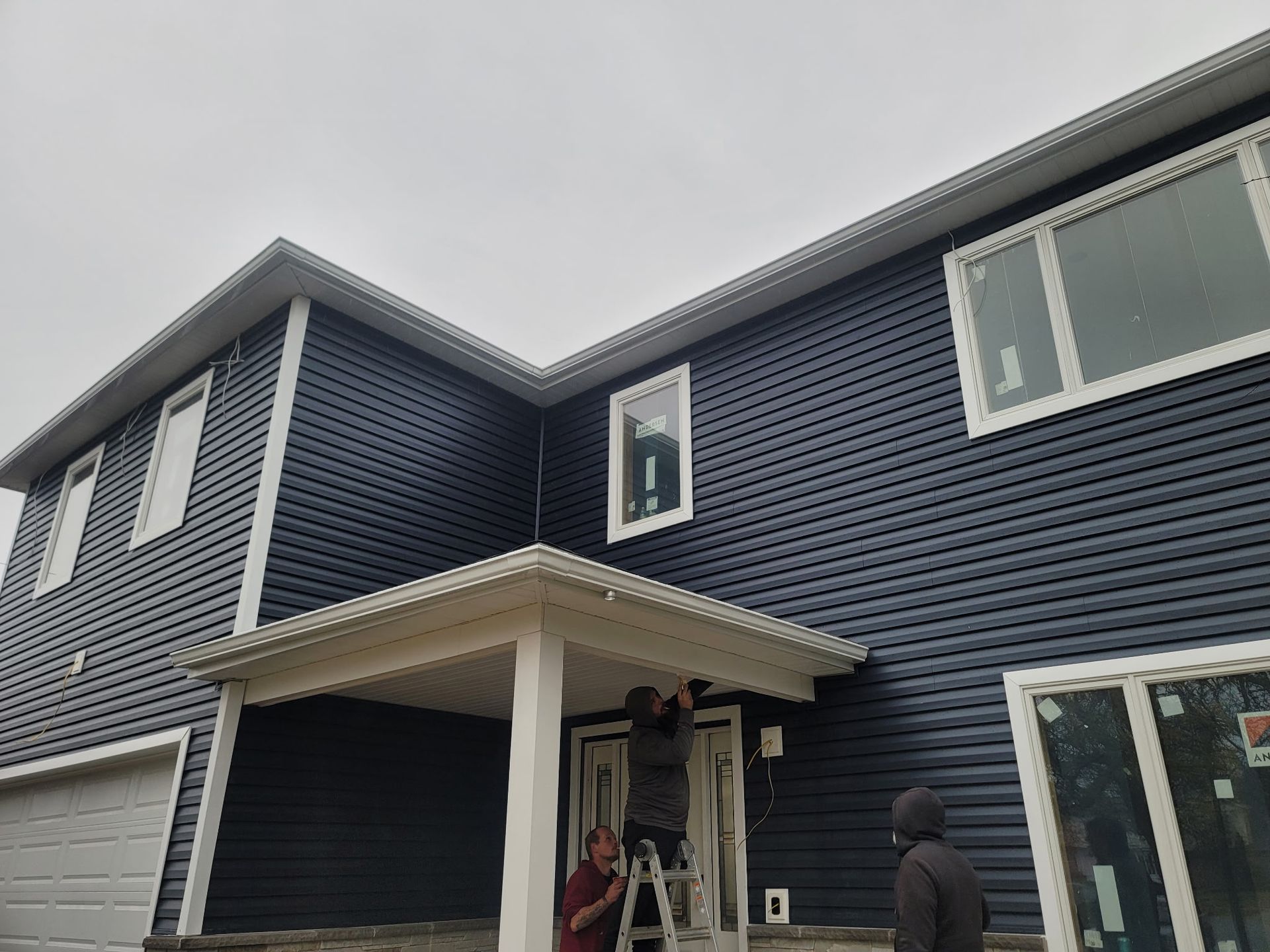 Two people work on installing trim above the front door of a two-story home with dark blue siding and a white porch.