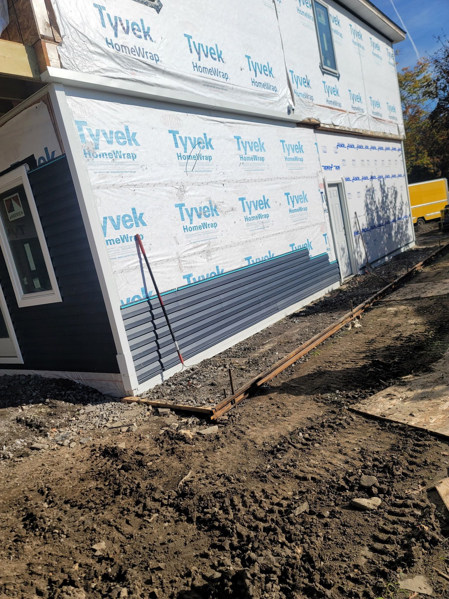Roofer on a roof, inspecting tiles. He wears work clothes, a tool belt, and a hat. Sunny day, outdoors.