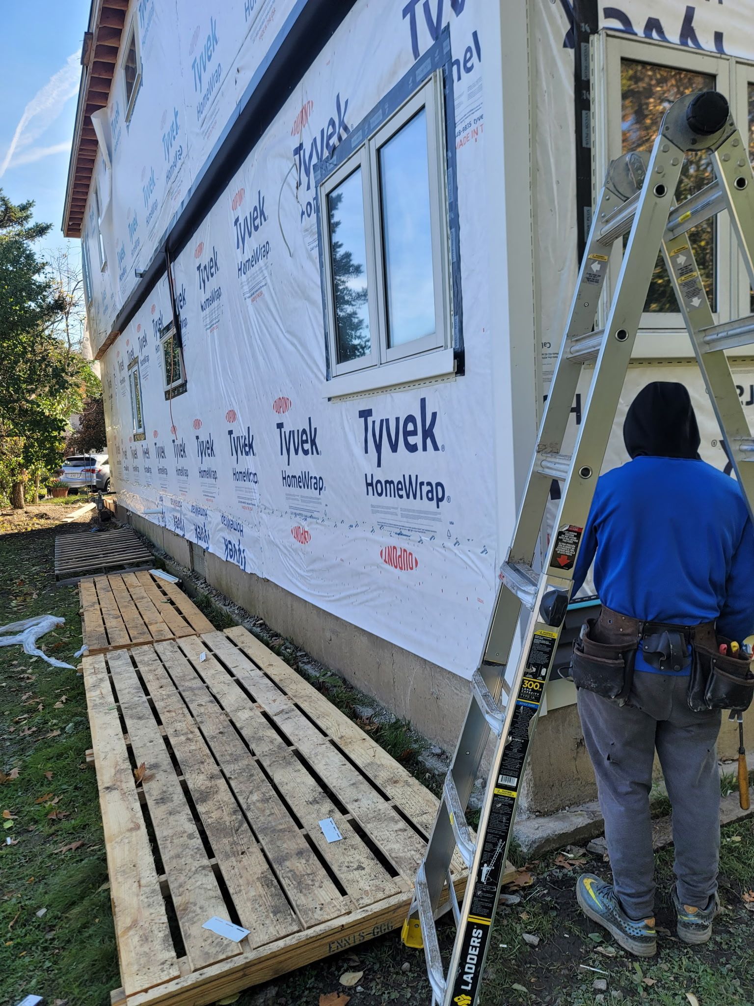 A person in a blue hoodie stands on a ladder near a house covered in white Tyvek building wrap and wooden pallets.