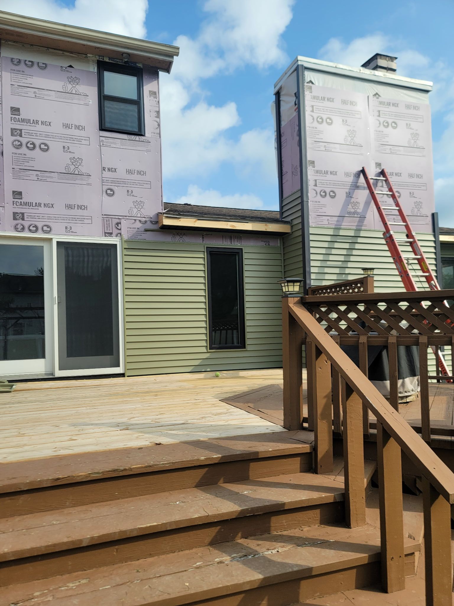 A house exterior under construction with exposed purple insulation board and light siding, featuring a deck and a ladder.