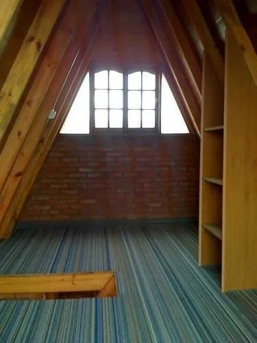 Habitación abuhardillada con techo inclinado, pared de ladrillo, ventana, estantes empotrados y alfombra azul a rayas.