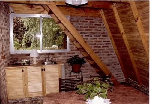 Habitación interior abuhardillada con ventana, pared de ladrillo, armarios de madera y una planta.
