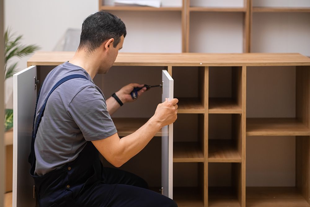 A man is sitting on the floor fixing a cabinet with a screwdriver.
