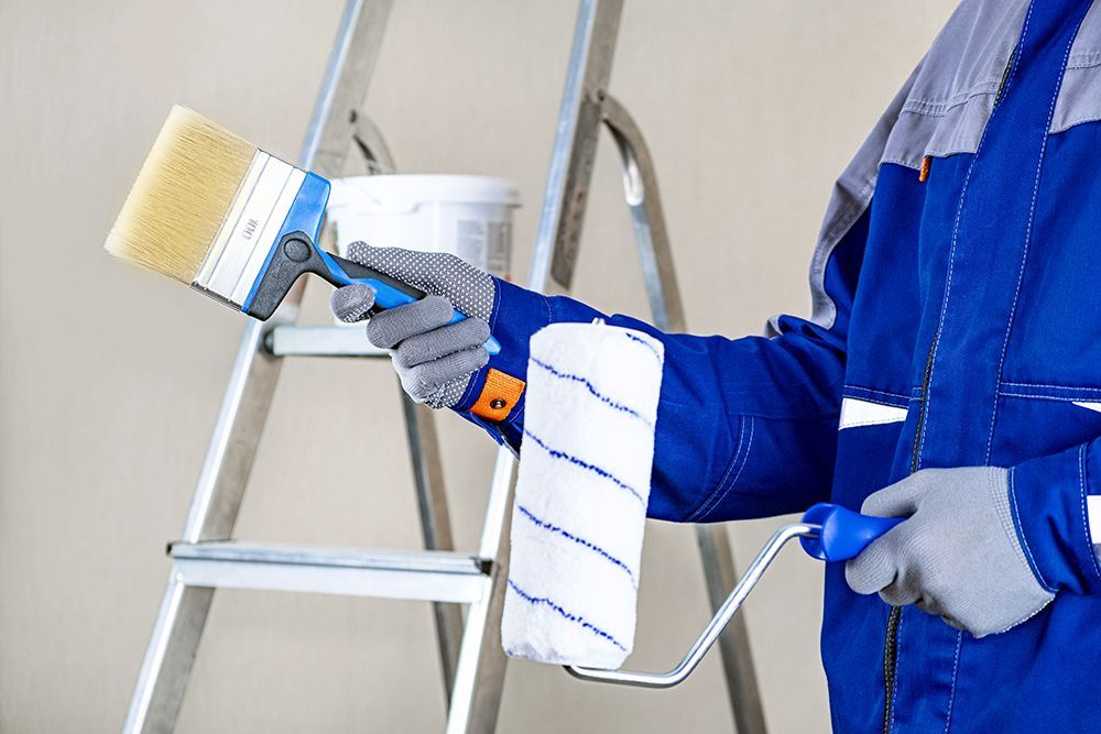 A person is holding a paint roller and a brush in front of a ladder.