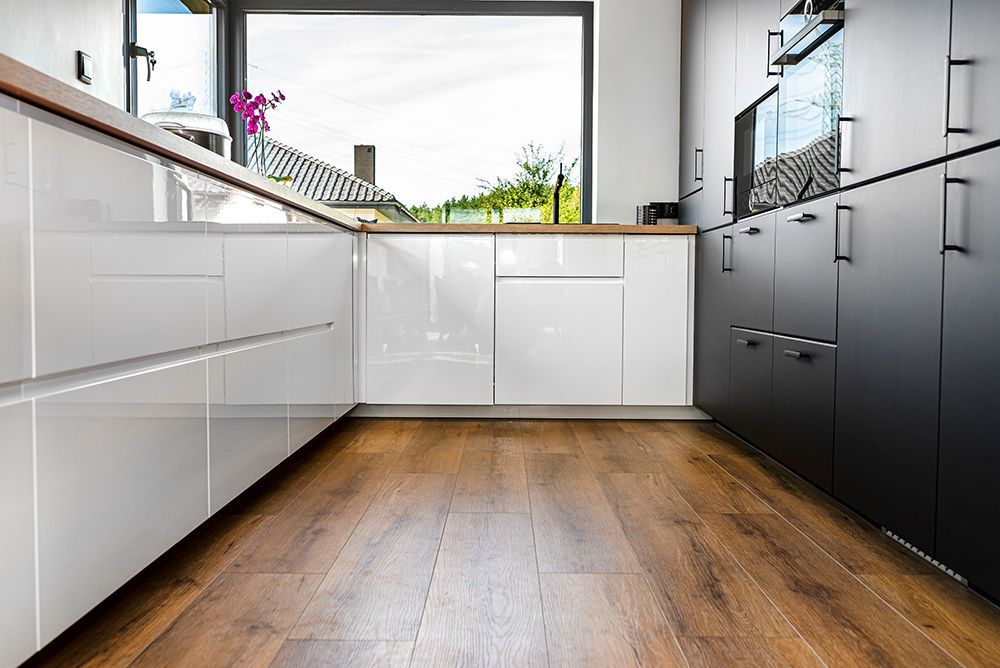 A kitchen with wooden floors , white cabinets and a large window.